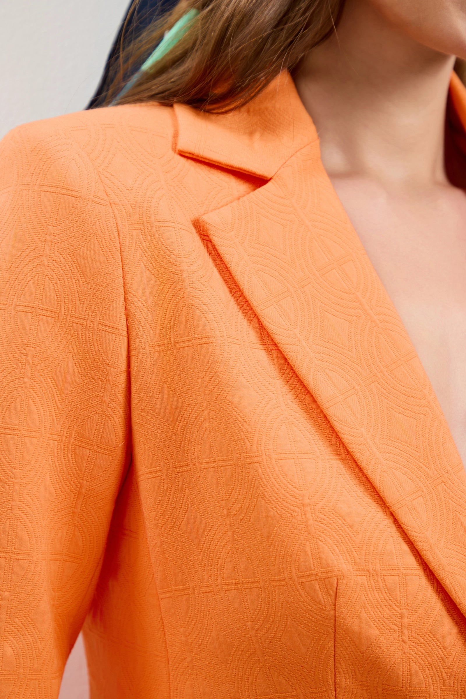 Close-up of a woman wearing the Incanta Fitted Blazer with an orange geometric texture, highlighting the lapel and shoulder, part of her neck, and wavy brown hair.