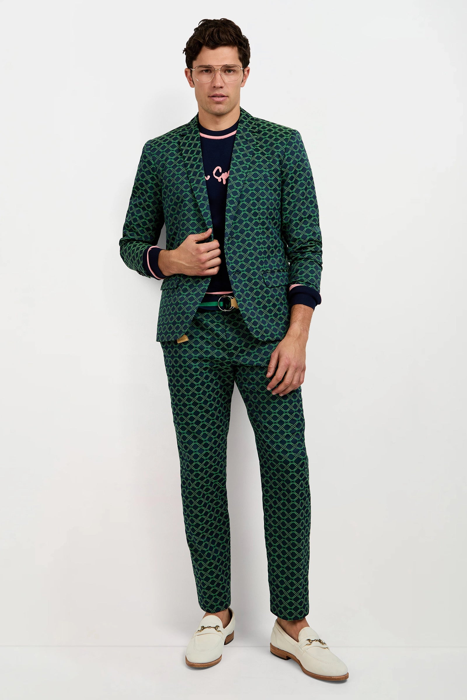 A man with short brown hair and glasses poses with one hand on his Thurston Blazer - Clover Geo Jacquard against a white background, styled with a black text shirt, black belt, and white loafers.