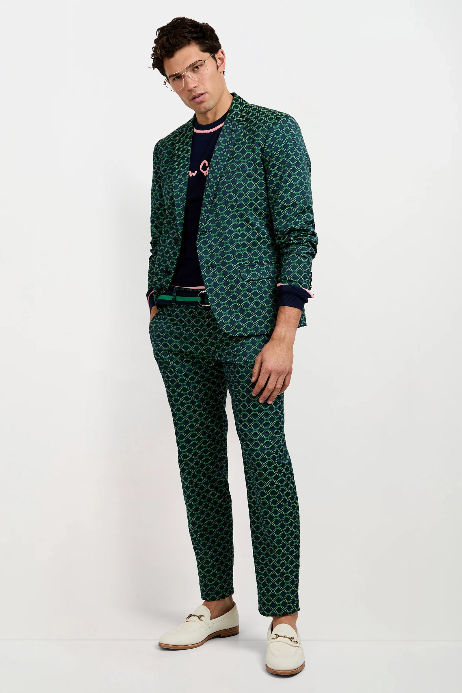 A man stands against a plain white background, one hand in his pocket, wearing glasses and the Clyde Slim Trouser in Clover Geo Jacquard, paired with a black shirt and white loafers.