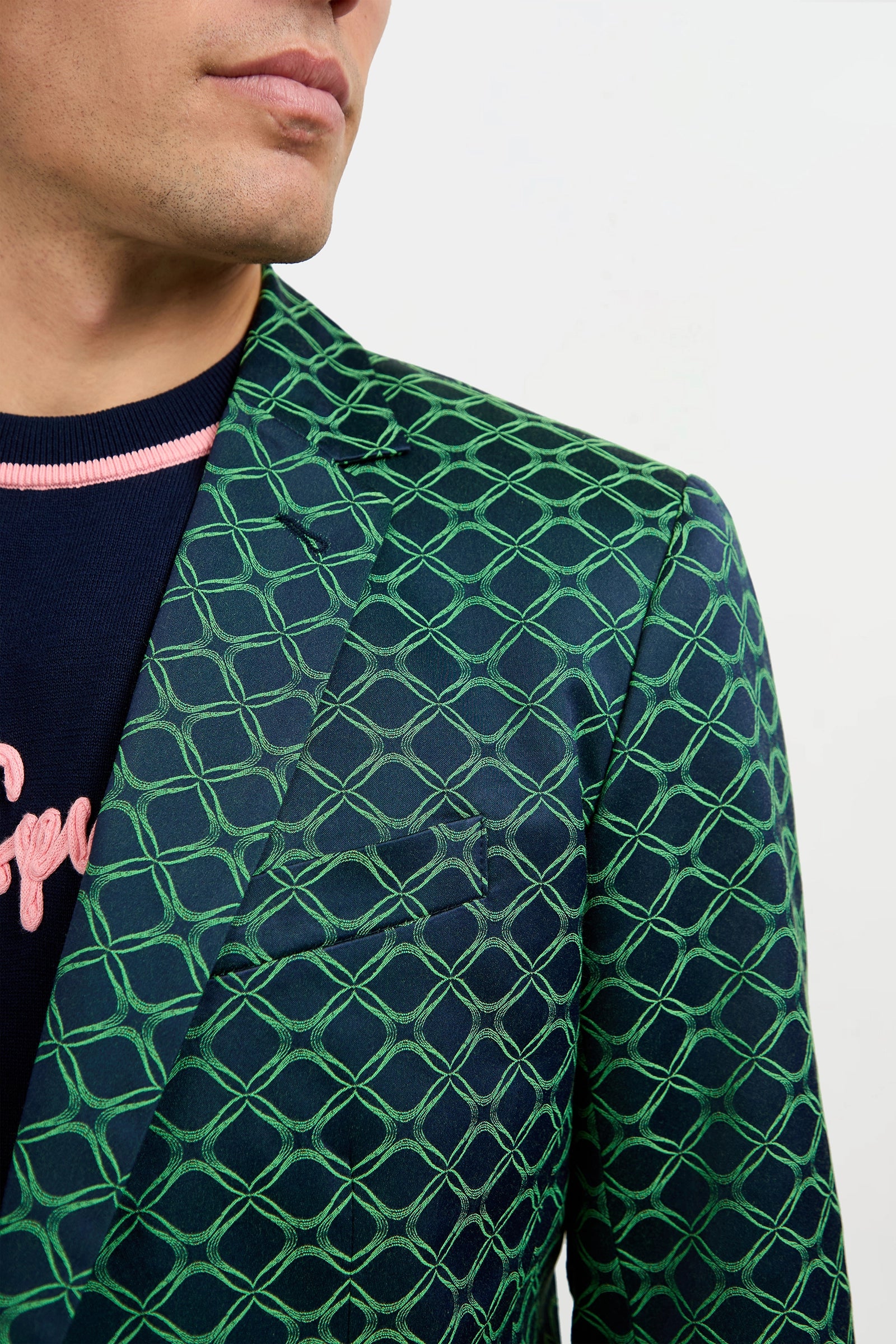 A man wears the Thurston Blazer in Clover Geo Jacquard—a dark blazer with a bold green geometric pattern—over a navy sweater with pink lettering, shown from the shoulders up against a plain white background.