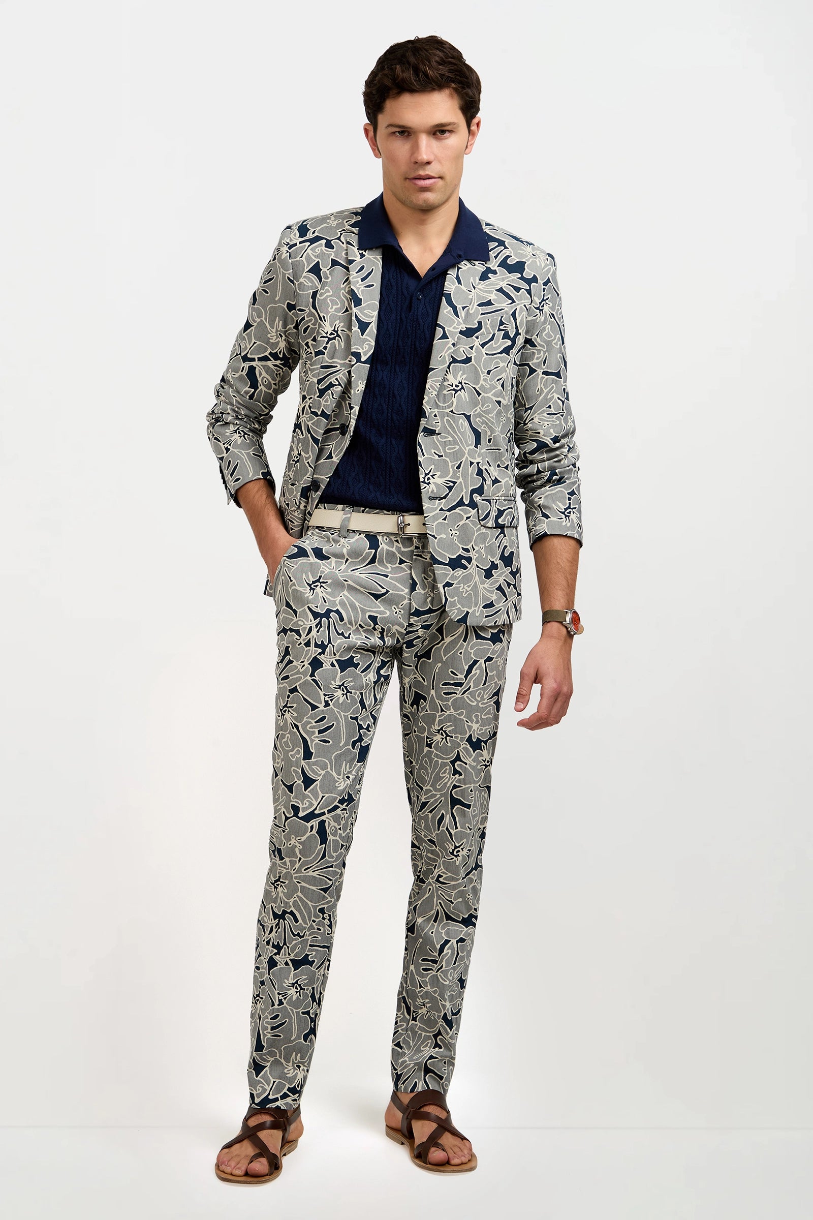 A man models the Thurston Blazer in Artsy Floral Jacquard, paired with a navy shirt and brown sandals. He stands against a white background with one hand in his pocket and a neutral expression.