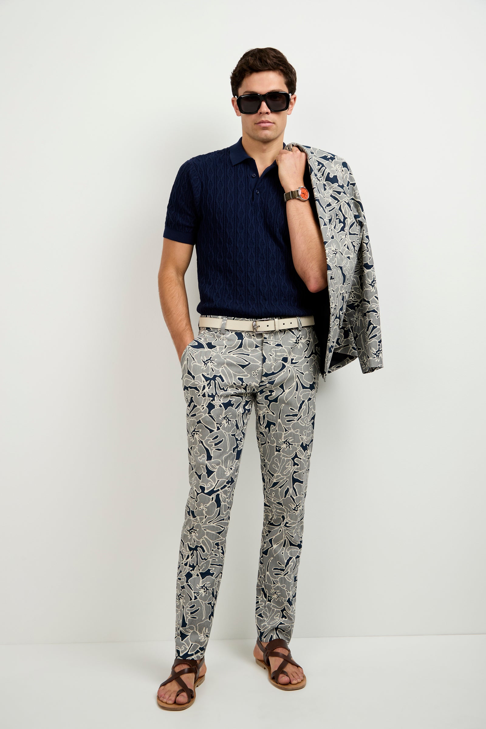 A man stands against a white wall wearing the Elio Short-Sleeve Polo, printed floral trousers, sunglasses, and brown sandals, with a matching floral jacket draped over his shoulder.