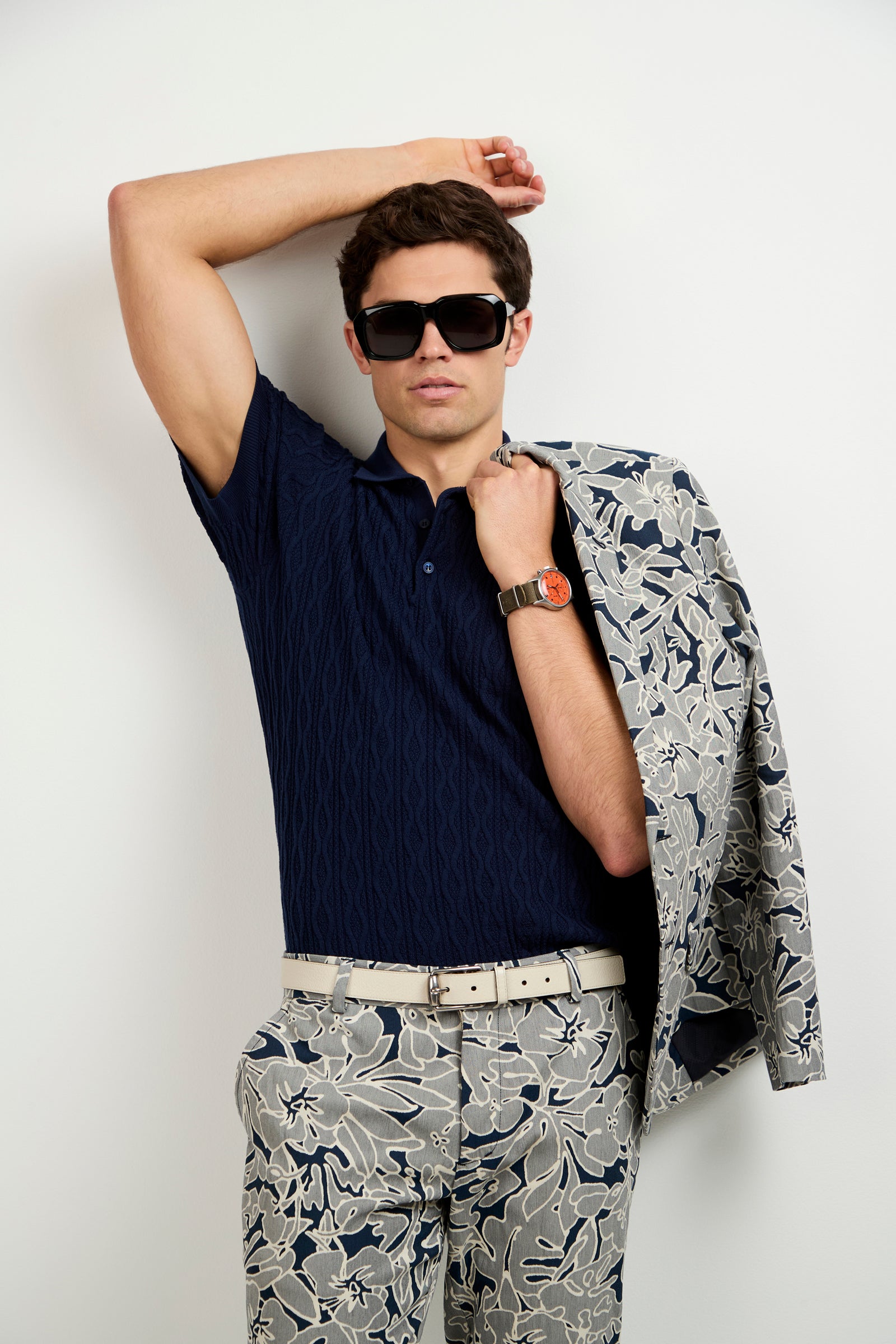 A man wearing the Elio Short-Sleeve Polo, dark sunglasses, floral-print pants, and a matching jacket draped over his shoulder poses against a plain light-colored wall.