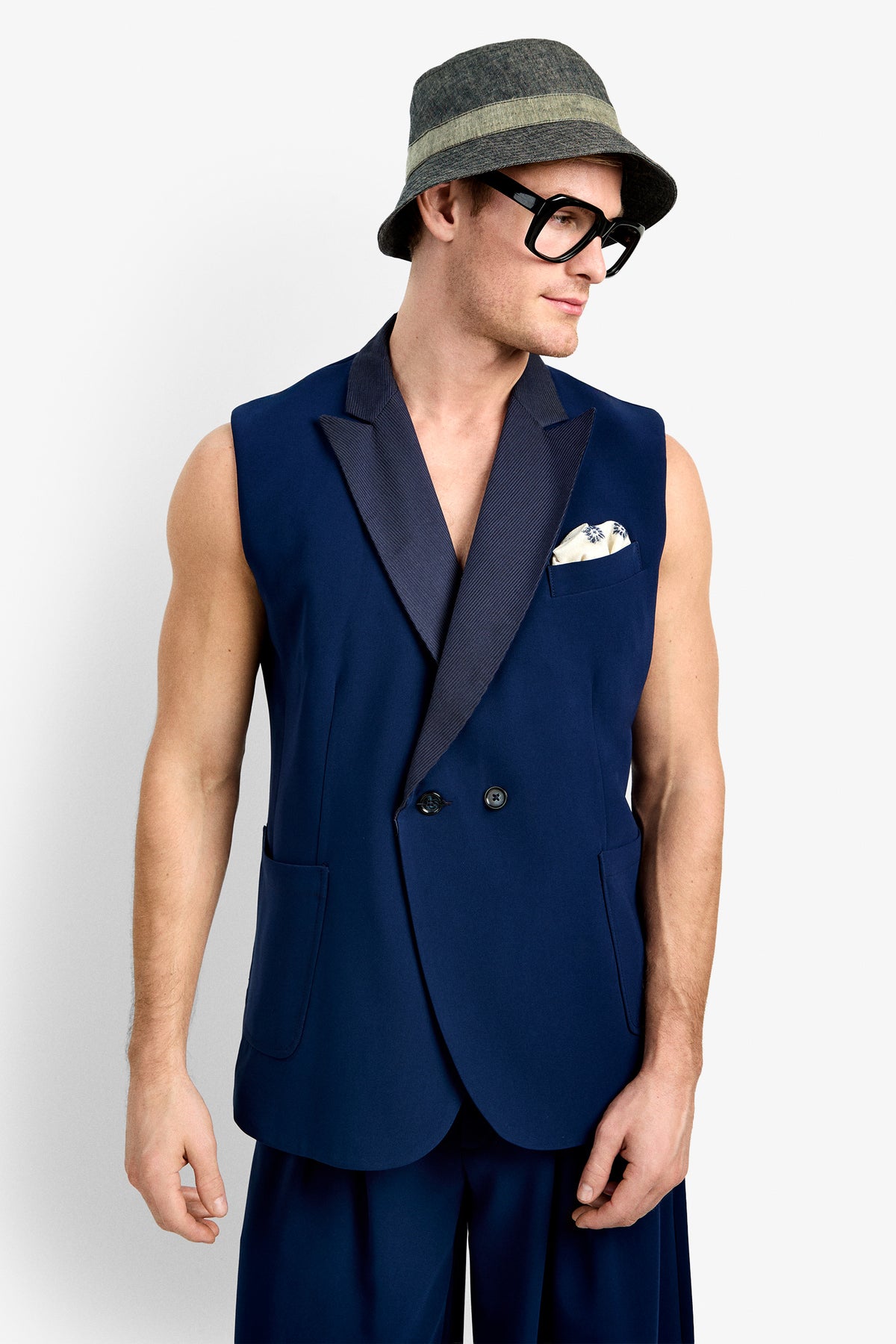 A man stands against a plain white background, looking to his left, wearing the Thurman Sleeveless Blazer with a dark lapel, a printed pocket square, wide navy pants, a gray-green bucket hat, and large black glasses.