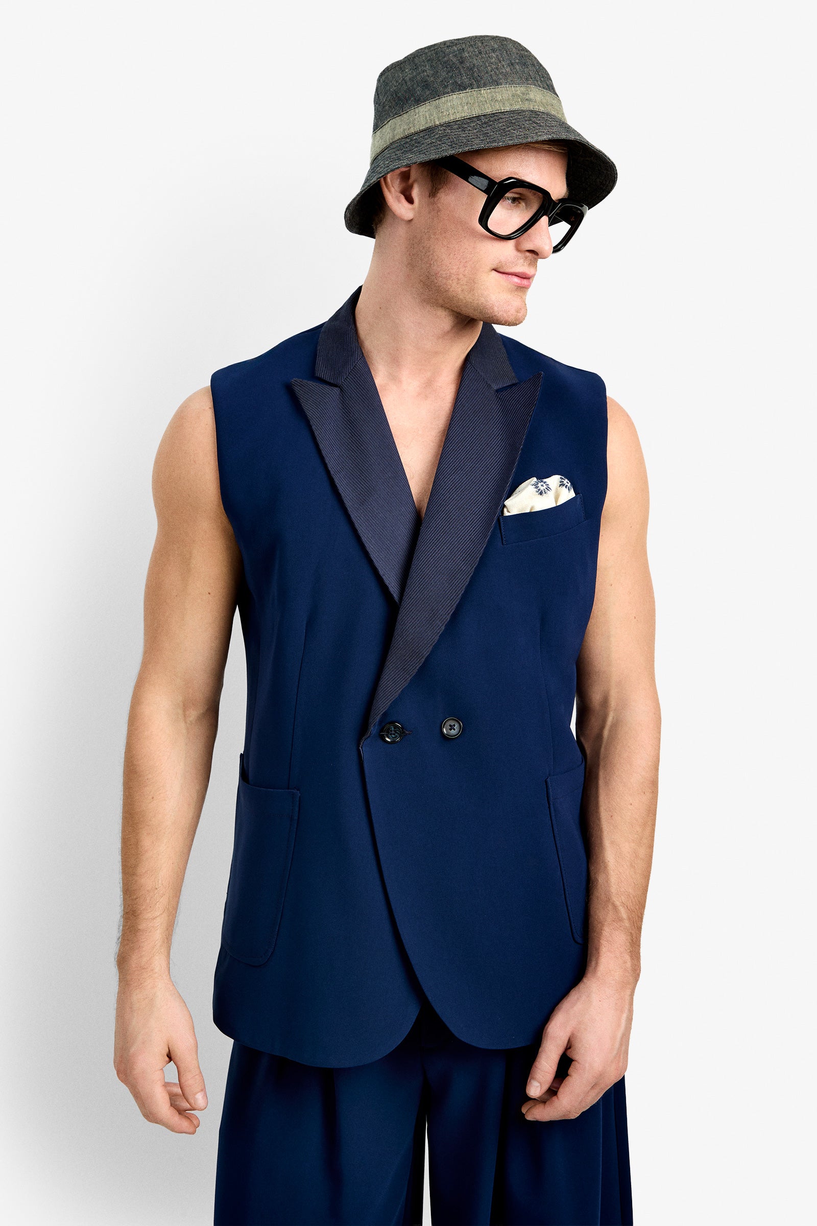 A man stands against a plain white background, looking to his left, wearing the Thurman Sleeveless Blazer with a dark lapel, a printed pocket square, wide navy pants, a gray-green bucket hat, and large black glasses.