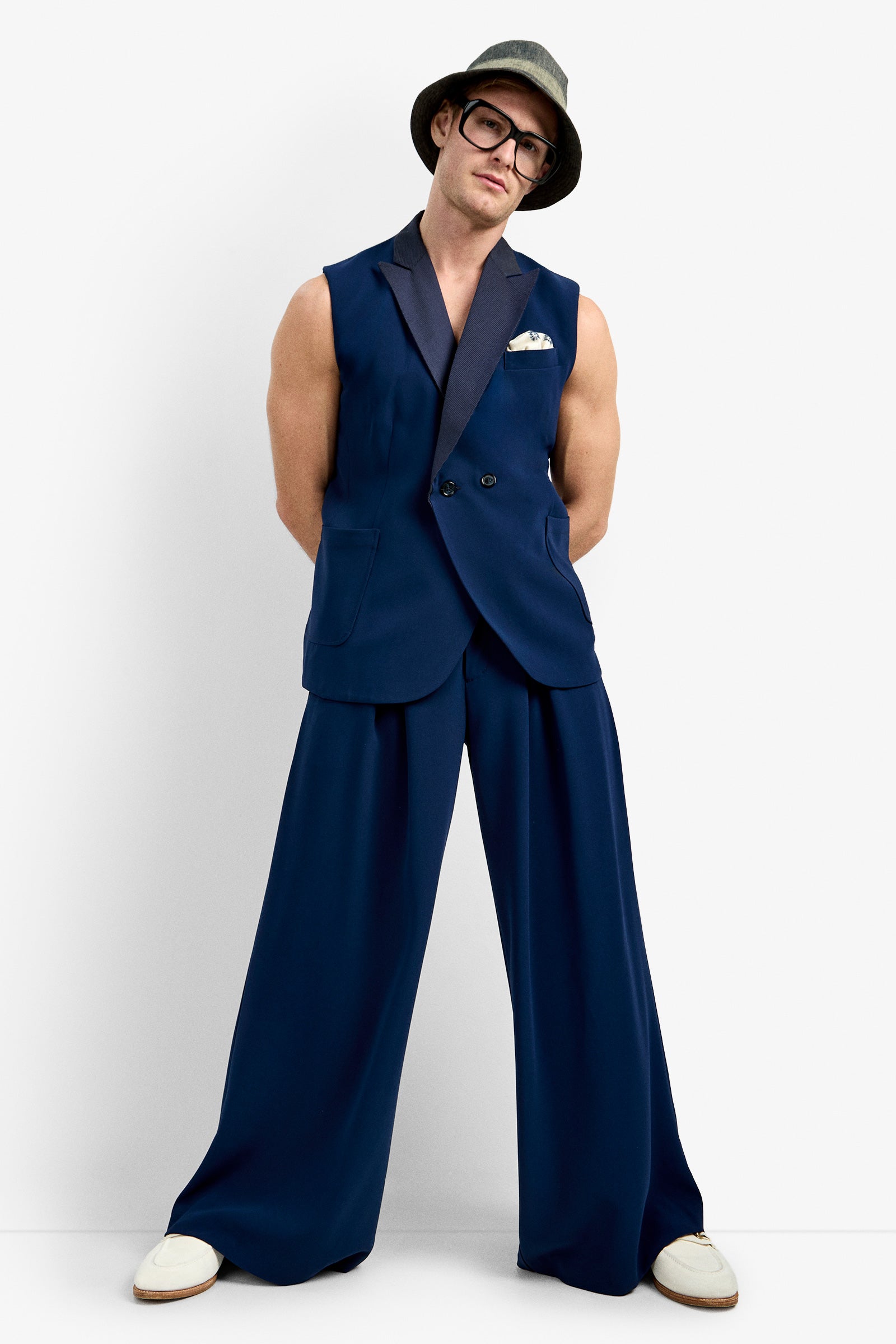 A person stands confidently against a plain white background wearing the Thurman Sleeveless Blazer in navy, wide-leg pants, white shoes, glasses, and a gray hat.