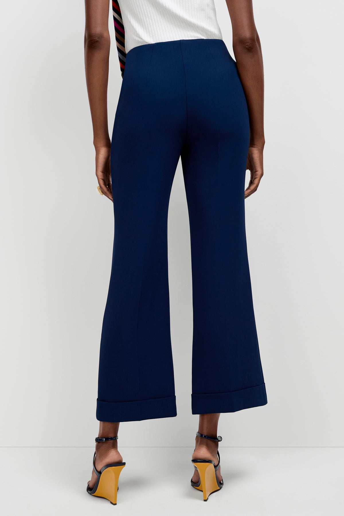 A person is seen from the back wearing Loren Cropped Kick-Flare Pants in dark blue with black heeled sandals featuring yellow soles, standing against a plain white background.