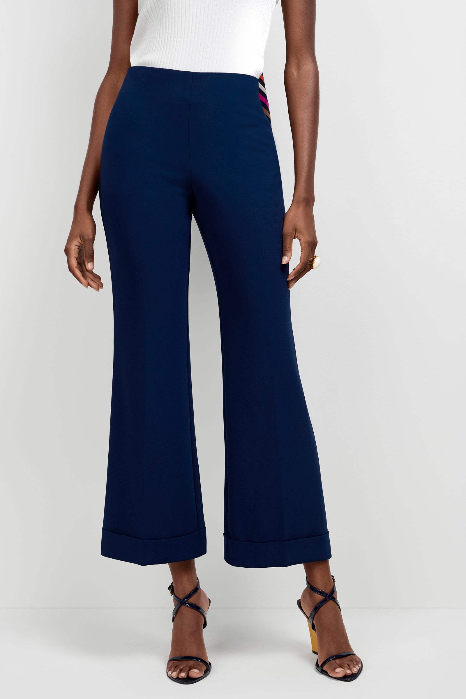 A person stands against a plain light background, wearing Loren Cropped Kick-Flare Pants in navy blue, paired with a white sleeveless top and strappy high-heeled sandals.