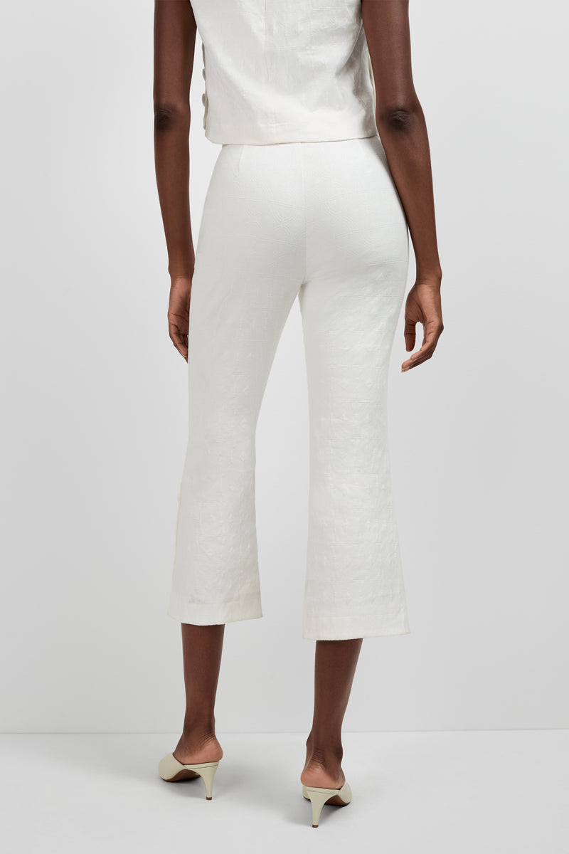 A person stands facing away, wearing Tortona Button Hem Pants in off-white, a matching sleeveless top, and cream-colored heeled shoes against a plain light background.