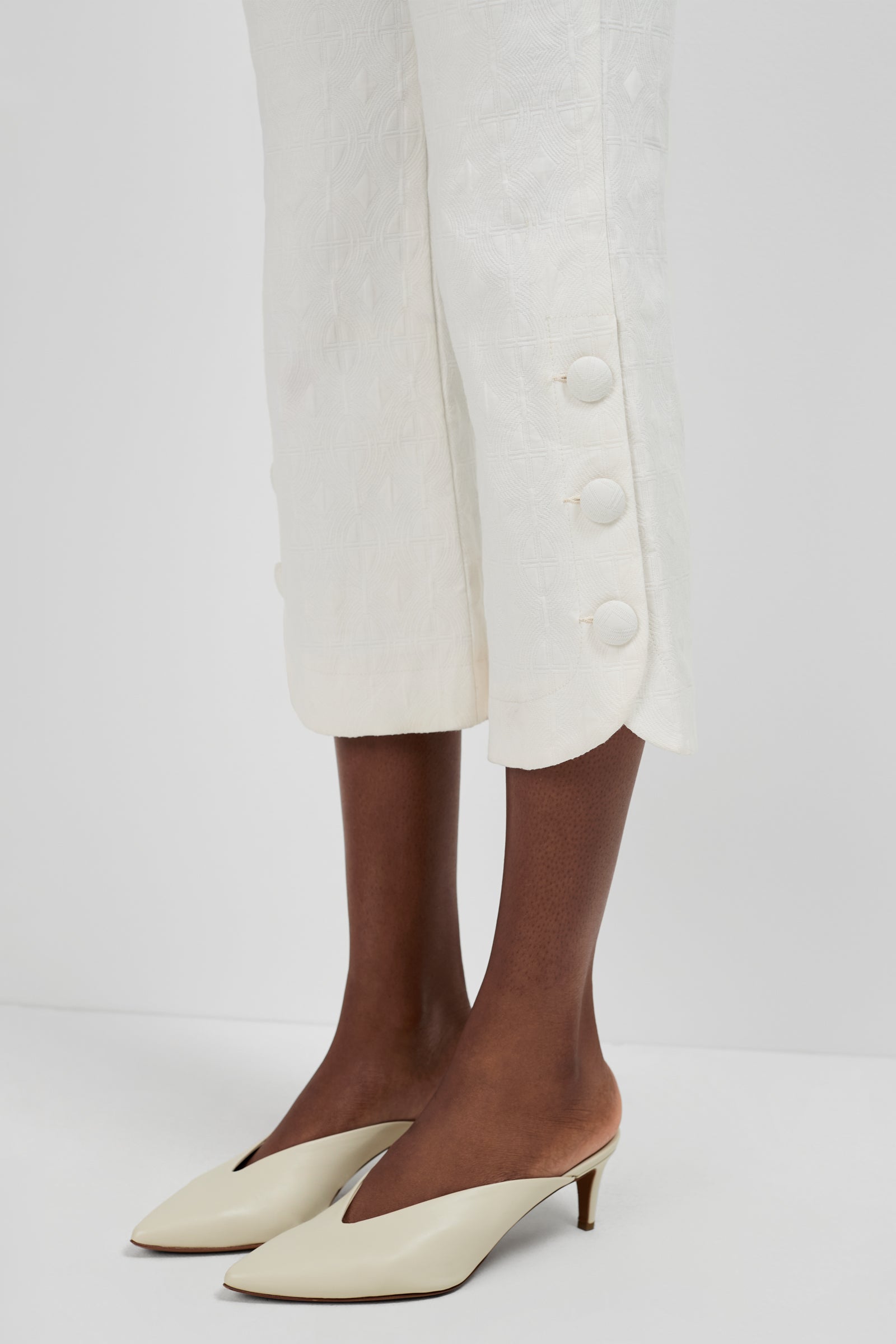 A person models the Tortona Button Hem Pants—white, textured, ankle-length with large decorative side buttons—paired with cream pointed-toe heeled mules, standing against a plain white background.