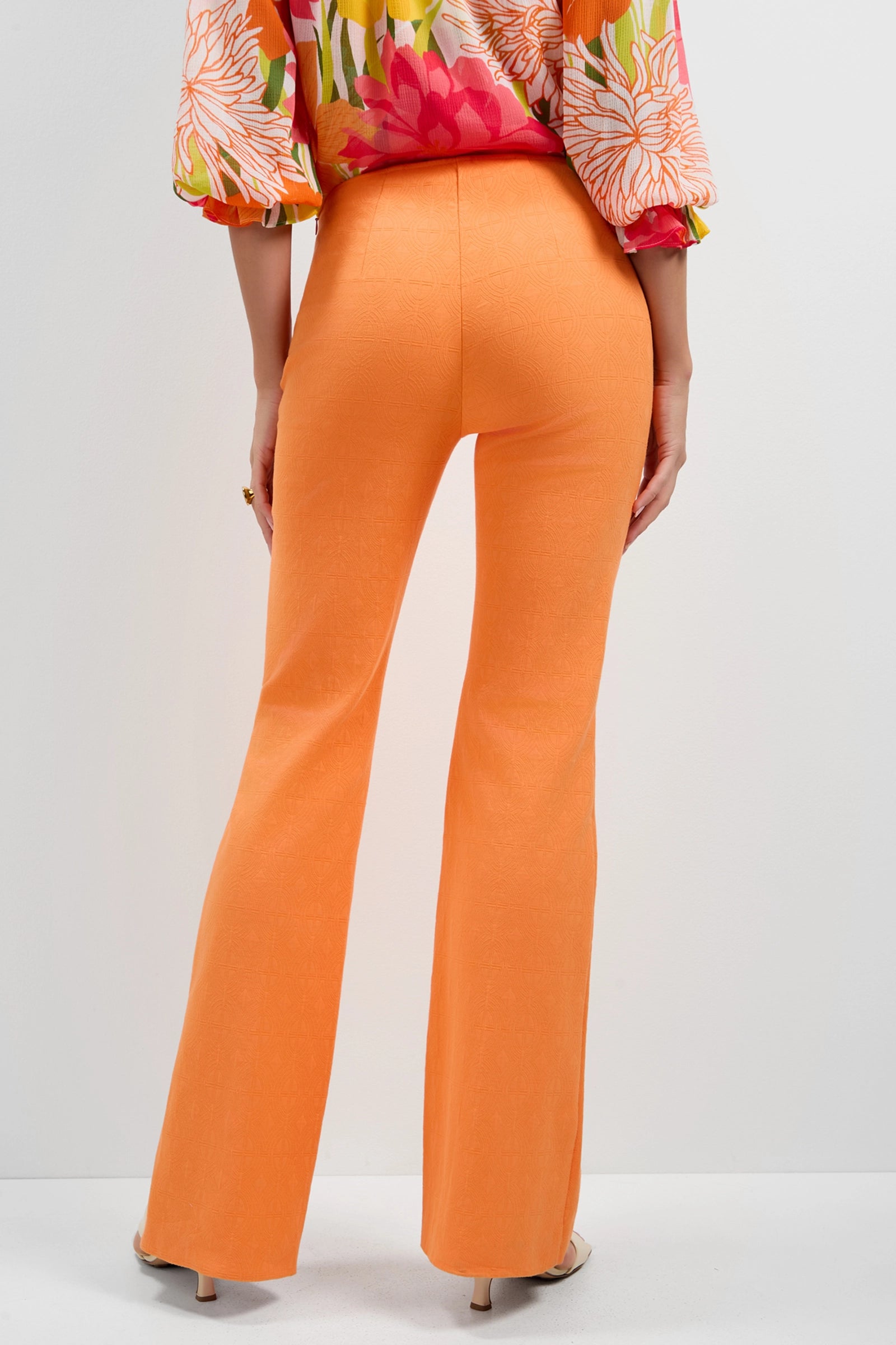 A person stands facing away in Jacoba Slim-Fit Bootcut Pants, bright orange and flared, paired with nude heels and a colorful floral long-sleeve blouse against a plain white background.