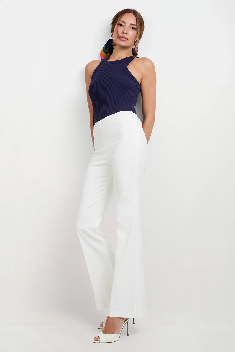 A woman stands against a white wall, wearing a navy blue sleeveless top, Jacoba Slim-Fit Bootcut Pants in white, white high-heeled shoes, and a colorful scarf tied in her hair.