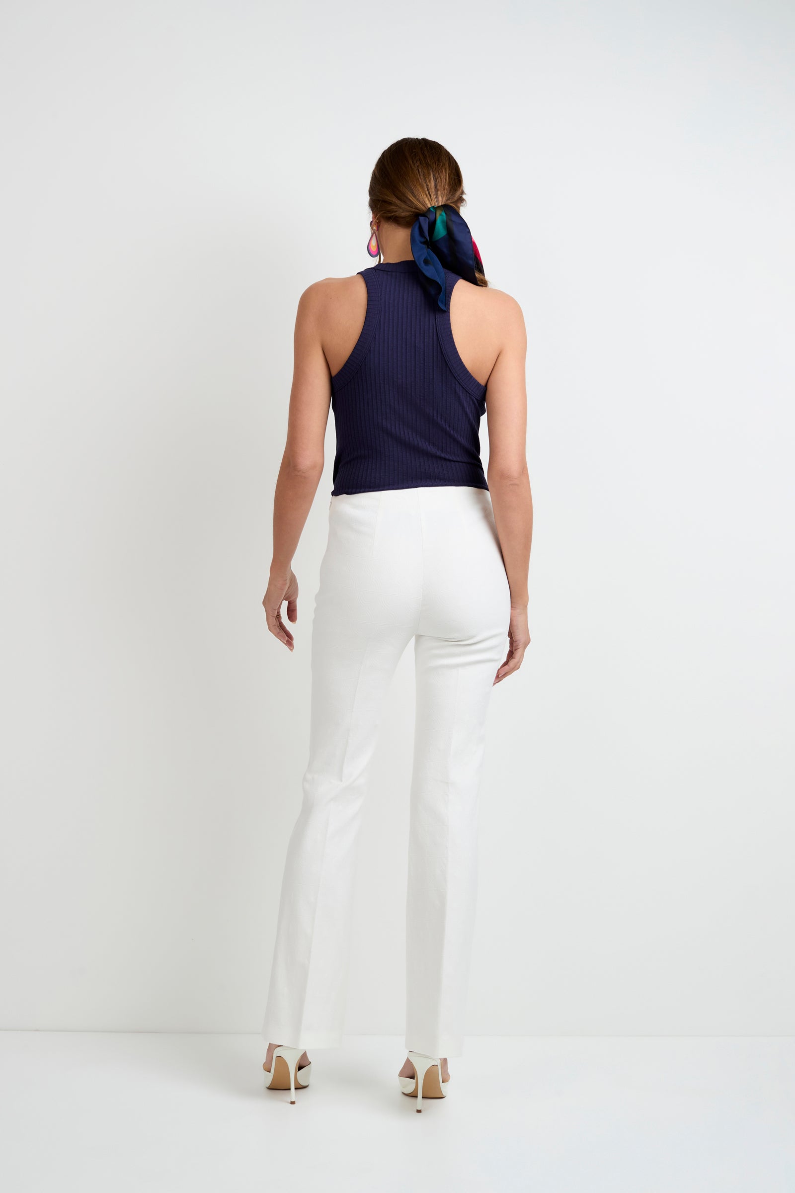 A woman faces away from the camera, wearing the Continental Tank Top in navy, white pants, white heels, and a colorful scarf in her hair, set against a plain white background.