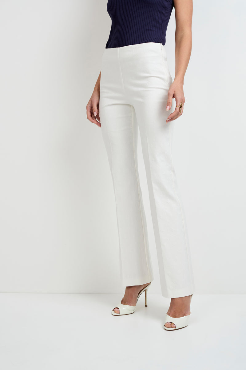 A woman stands against a white background wearing Jacoba Slim-Fit Bootcut Pants in white, a sleeveless navy top, and white heeled sandals. Her arms are relaxed by her sides.