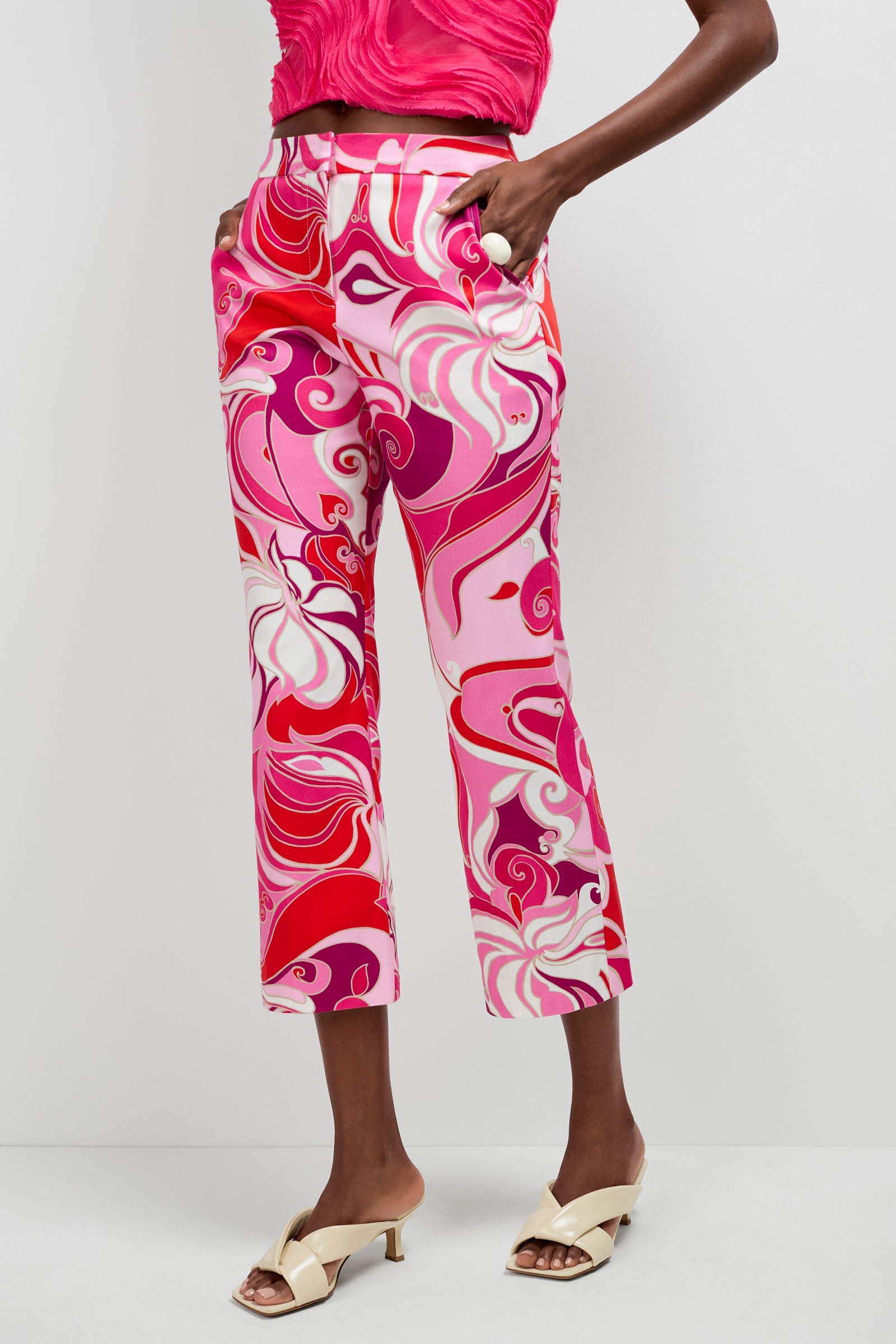 Someone is wearing the Lulu Cropped Pants - Darsana, which feature pink, red, and white swirls, along with a pink textured crop top and cream open-toe heeled sandals, standing against a plain white background.