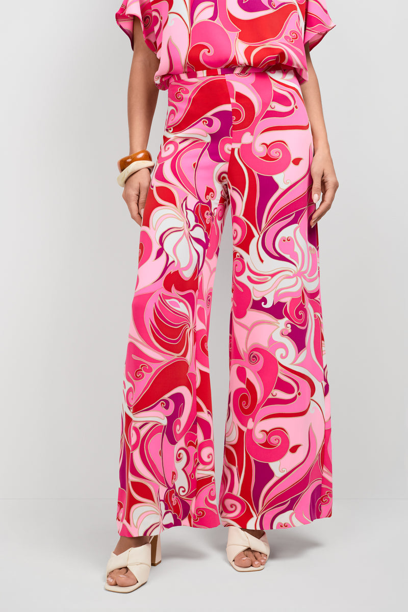 Someone models the Long Weekend Pants, featuring a bold, swirling pink, red, and white abstract print. Styled with a matching top, white open-toe heels, and chunky bracelets, they stand against a plain background.