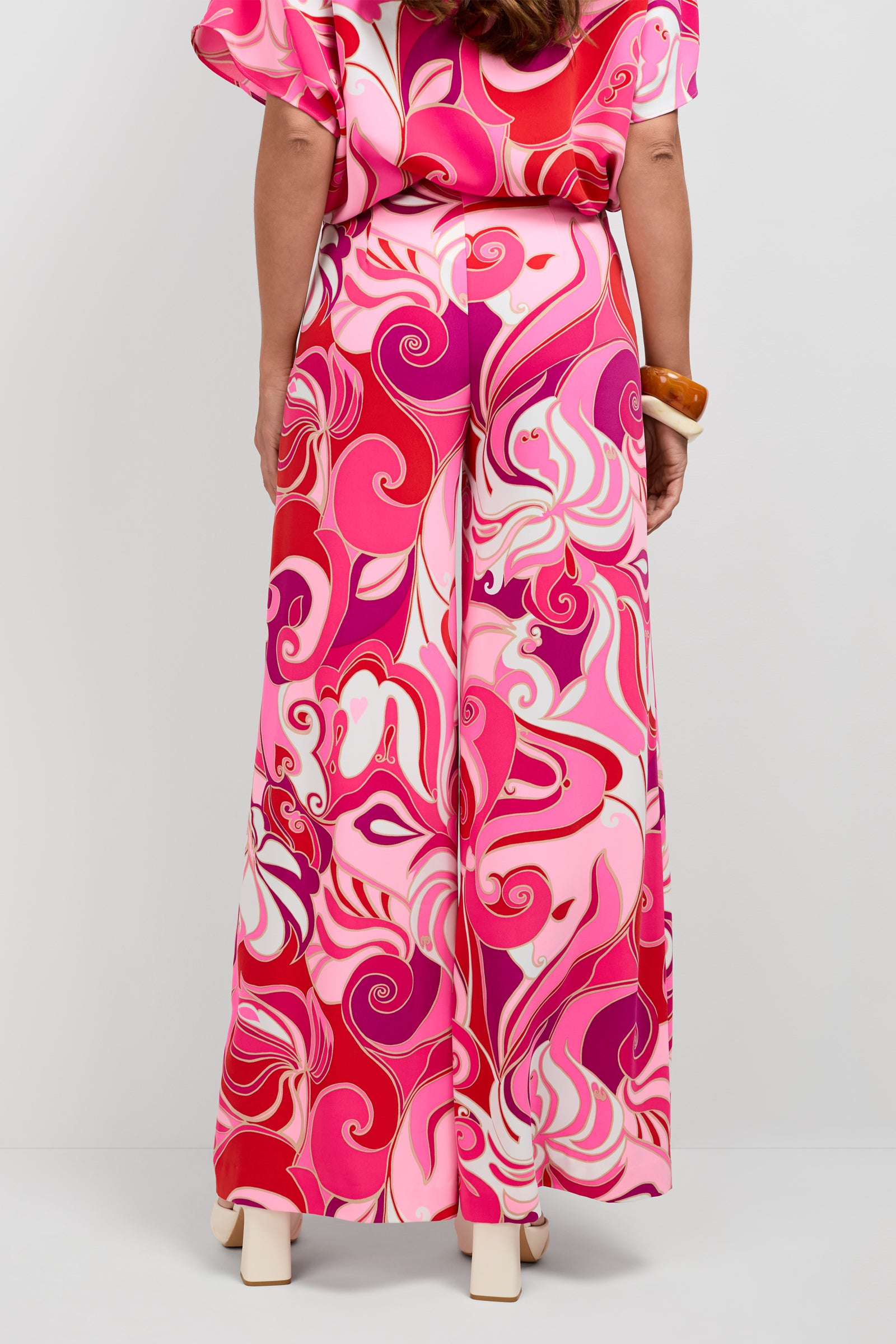 A person stands facing away, wearing Long Weekend Pants with a pink, red, and white swirl pattern, a matching top, cream platform heels, and a chunky brown bracelet on their right wrist.