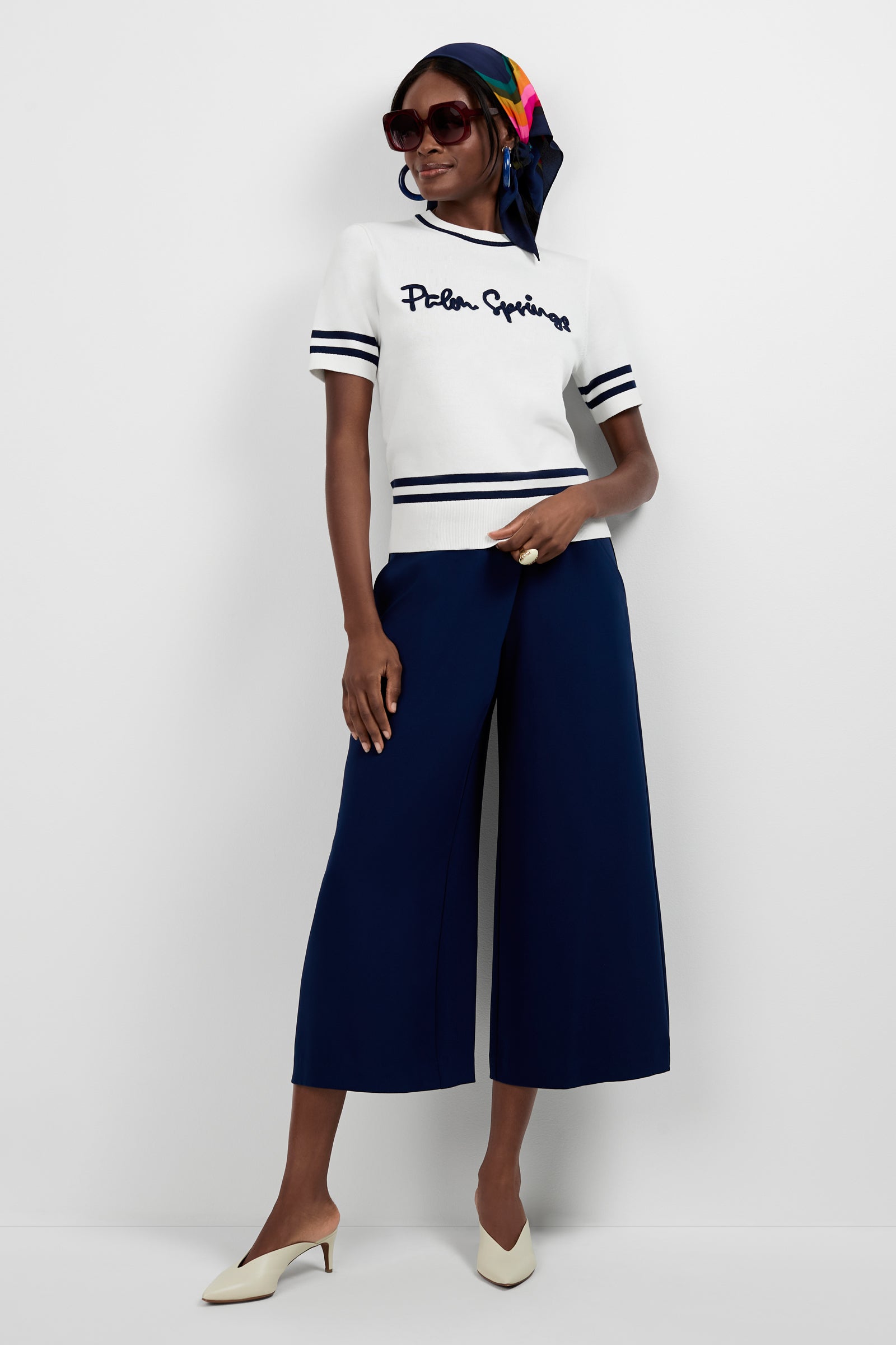 A woman stands against a white background wearing the “Old Fashioned Sweater - Palm Springs,” navy wide-leg pants, cream heels, dark sunglasses, and a colorful scarf tied over her hair.