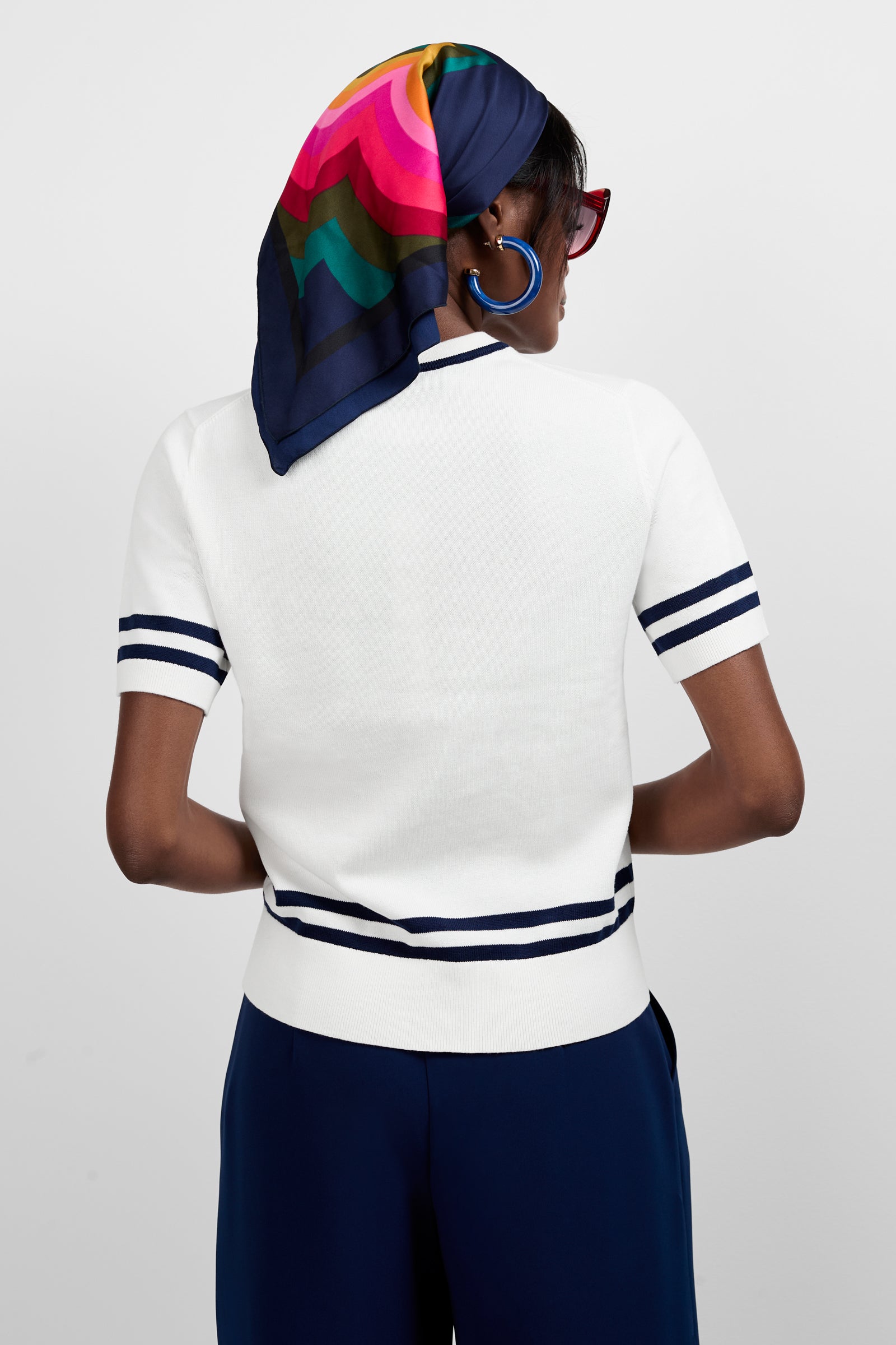 A person stands facing away, wearing the Old Fashioned Sweater - Palm Springs, a white short-sleeve sweater with navy stripes on the sleeves and hem, paired with navy pants, a colorful headscarf, bold blue hoops, and red sunglasses.