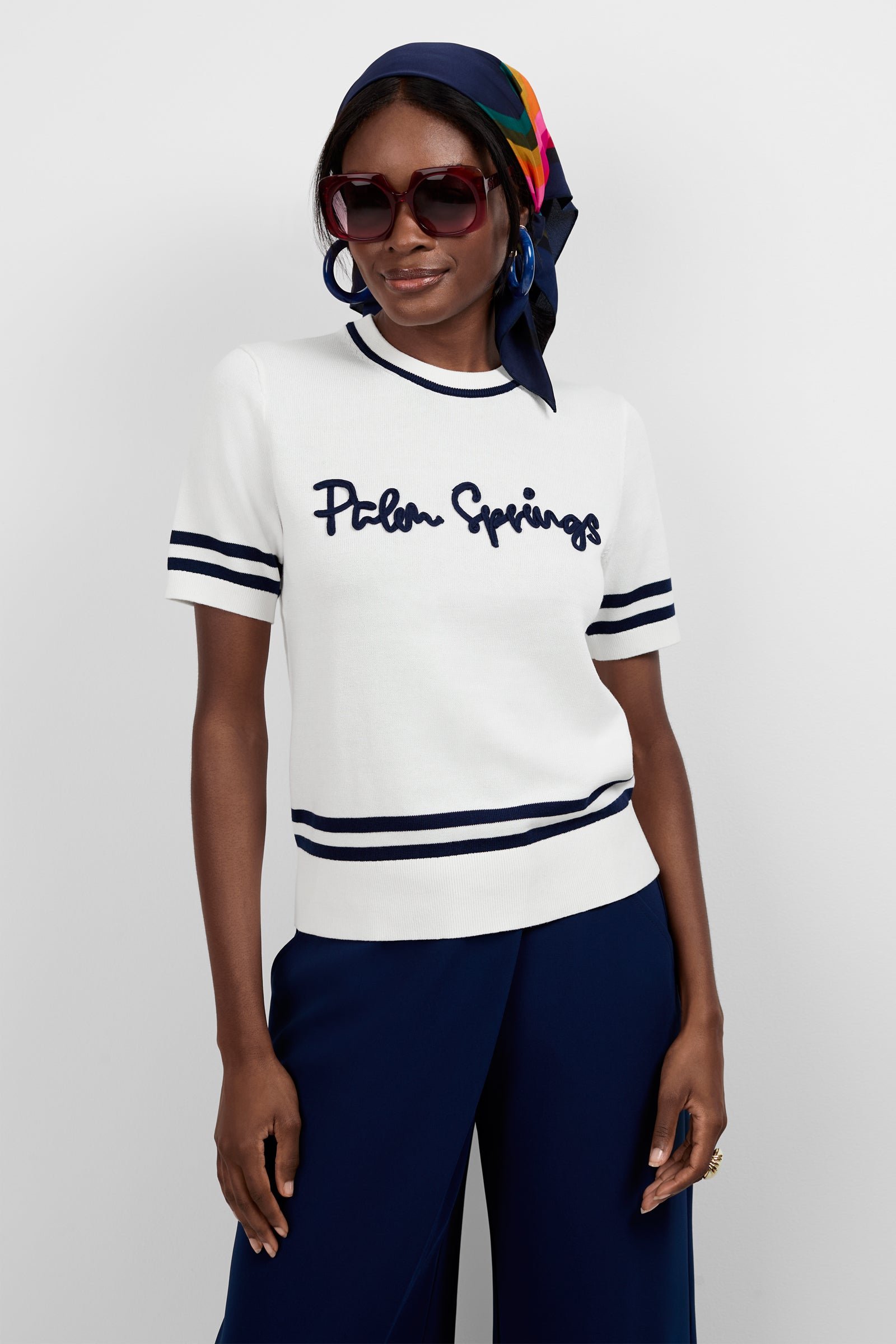 A woman models the Old Fashioned Sweater - Palm Springs, featuring navy stripes and a Palm Springs print, styled with sunglasses, a colorful headscarf, blue earrings, and navy blue pants against a plain background.