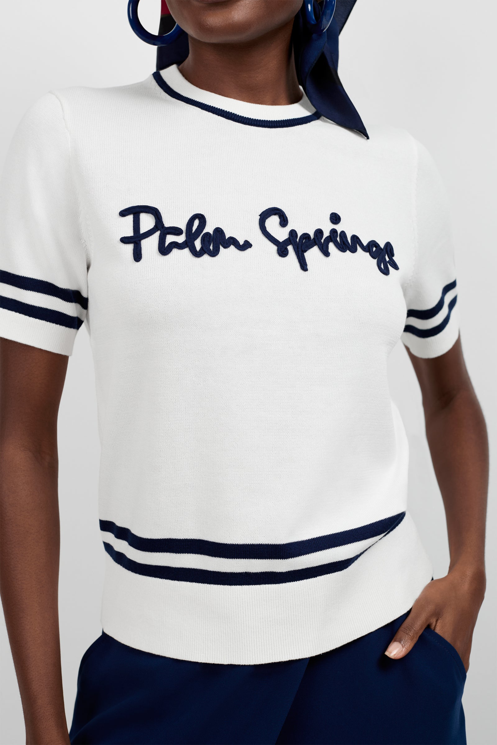 A woman wears the Old Fashioned Sweater - Palm Springs, a white short-sleeve sweater featuring navy stripes on the sleeves, collar, and hem, with "Palm Springs" in navy script across the chest.