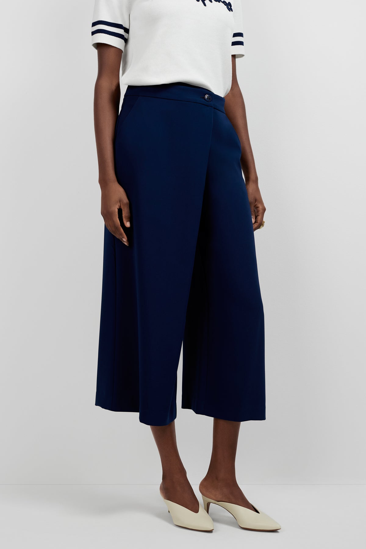 A person wearing Conchetta Wide-Leg Pants in navy blue, a white short-sleeve shirt with dark sleeve stripes, and cream low-heeled shoes, standing against a plain light background.