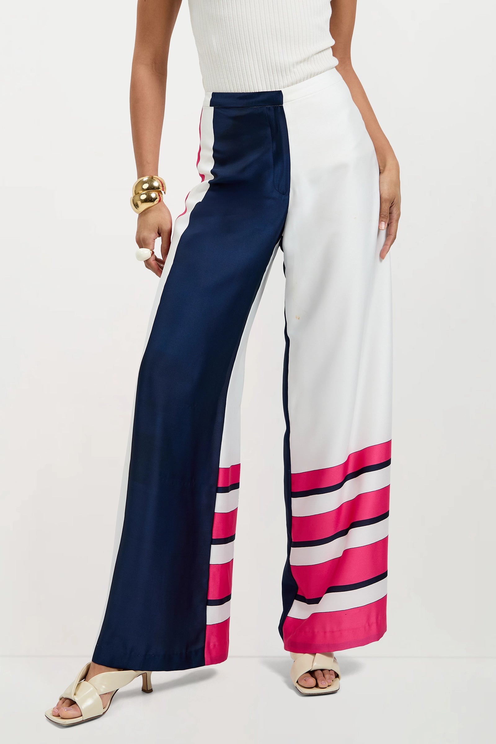 A woman wears Lanay Linee Wide-Leg Pants with one navy and one white leg, both featuring bold pink stripes near the hem, paired with a sleeveless white top and heeled sandals.