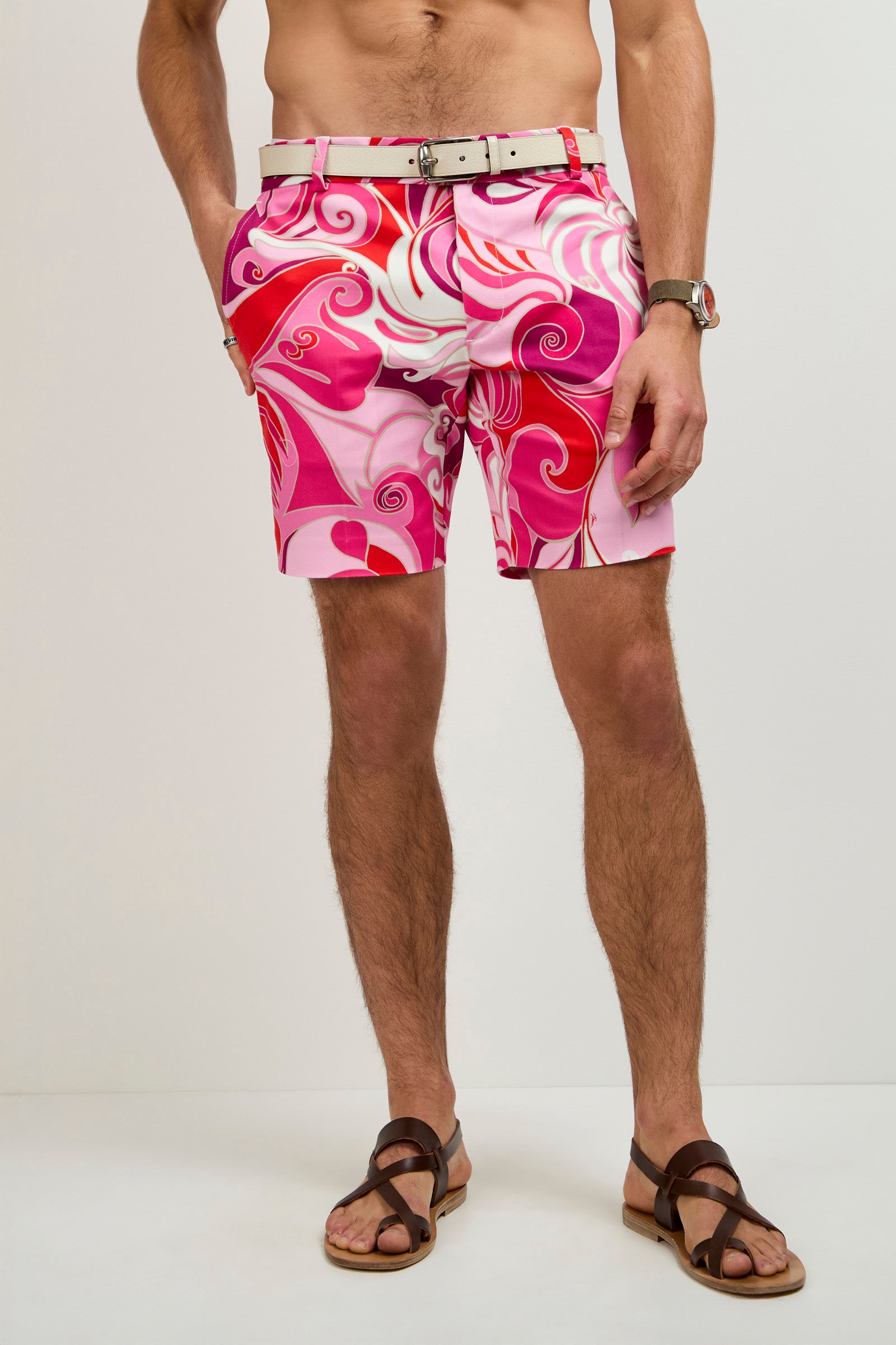 A man models the Lawrence Short - Darsena, featuring vibrant pink and red swirls, paired with a white belt, brown sandals, and a wristwatch. His upper body is not shown as he stands against a plain background with hands in his pockets.