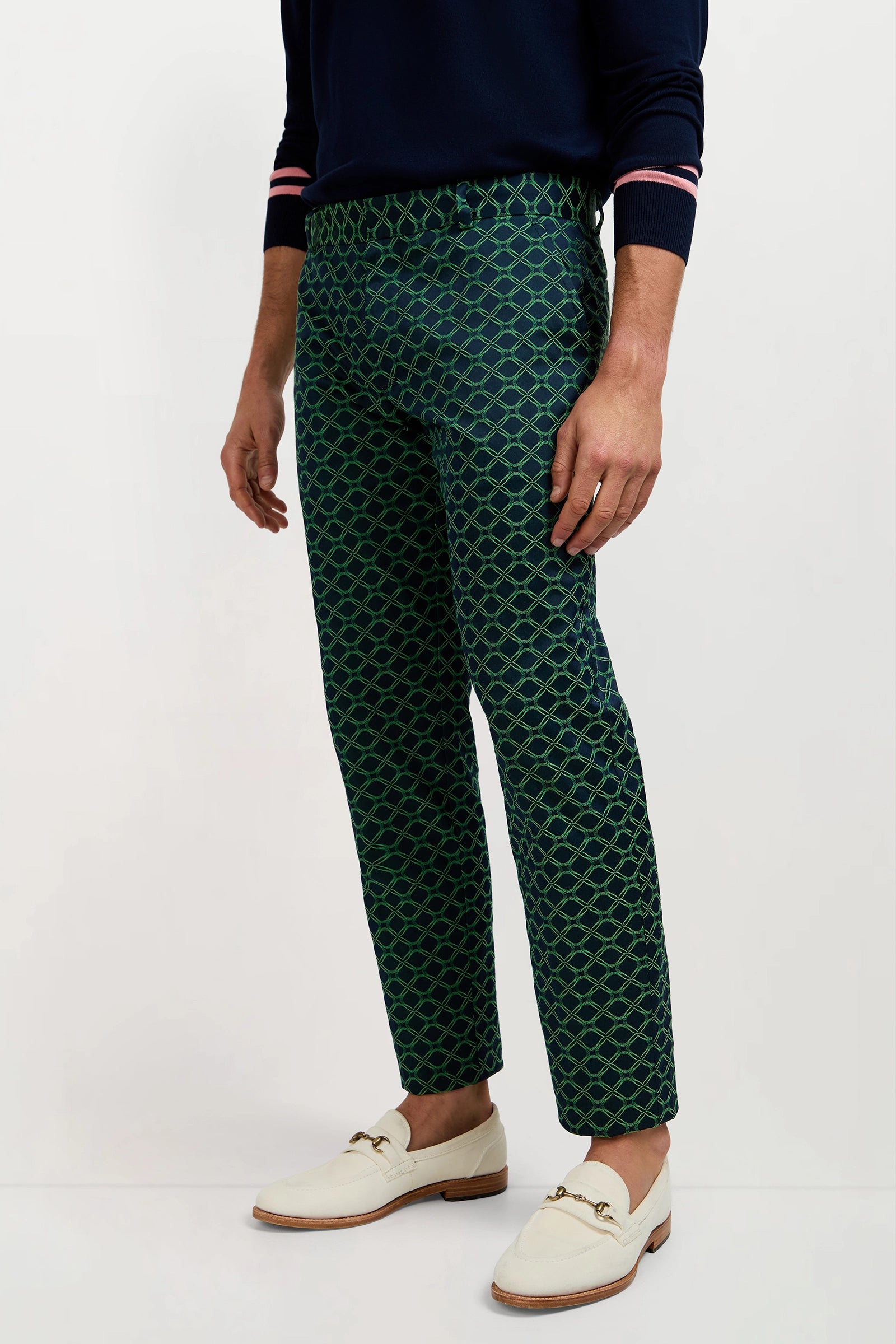 Wearing the Clyde Slim Trouser in Clover Geo Jacquard—patterned green and navy trousers—a person stands against a plain background, styled with a navy sweater and white loafers.