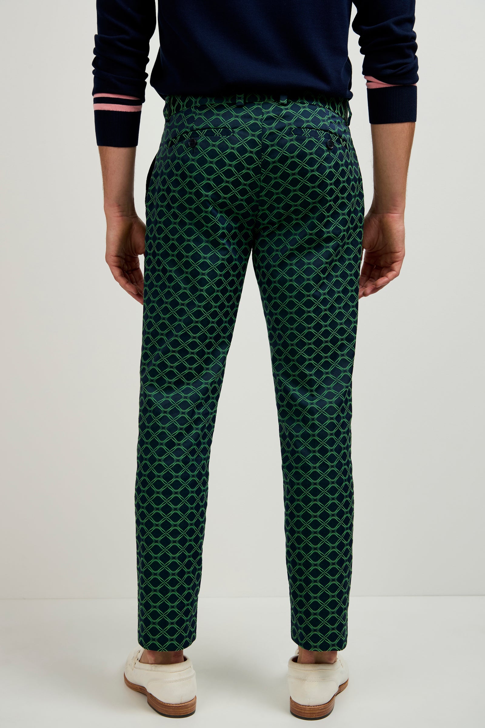 A person stands facing away, wearing the Clyde Slim Trouser - Clover Geo Jacquard with a navy sweater featuring pink-striped cuffs and white shoes, set against a plain, light-colored background.