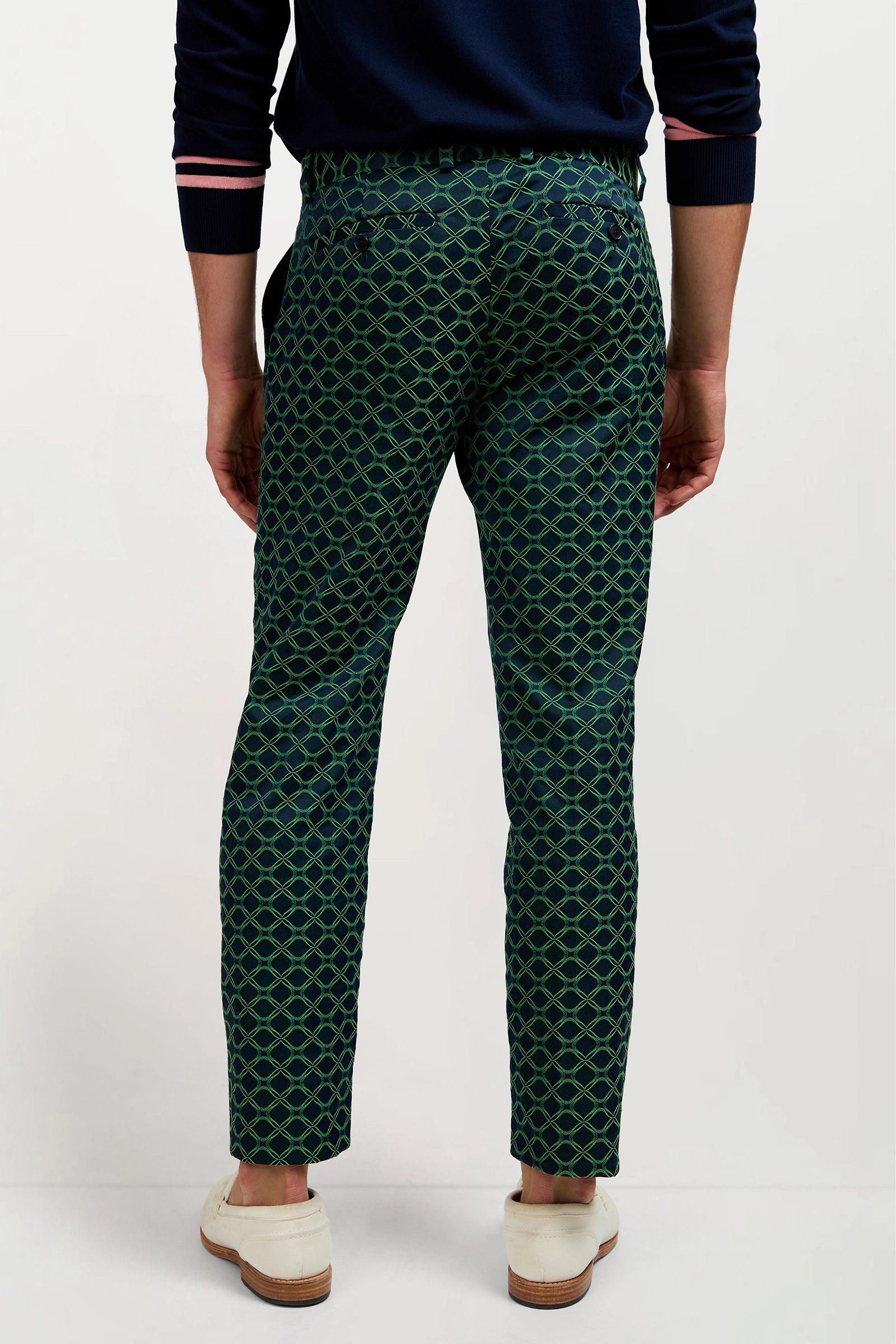 A person stands facing away, wearing the Clyde Slim Trouser in Clover Geo Jacquard—a dark green pant with a blue geometric pattern—paired with a navy sweater and white shoes.
