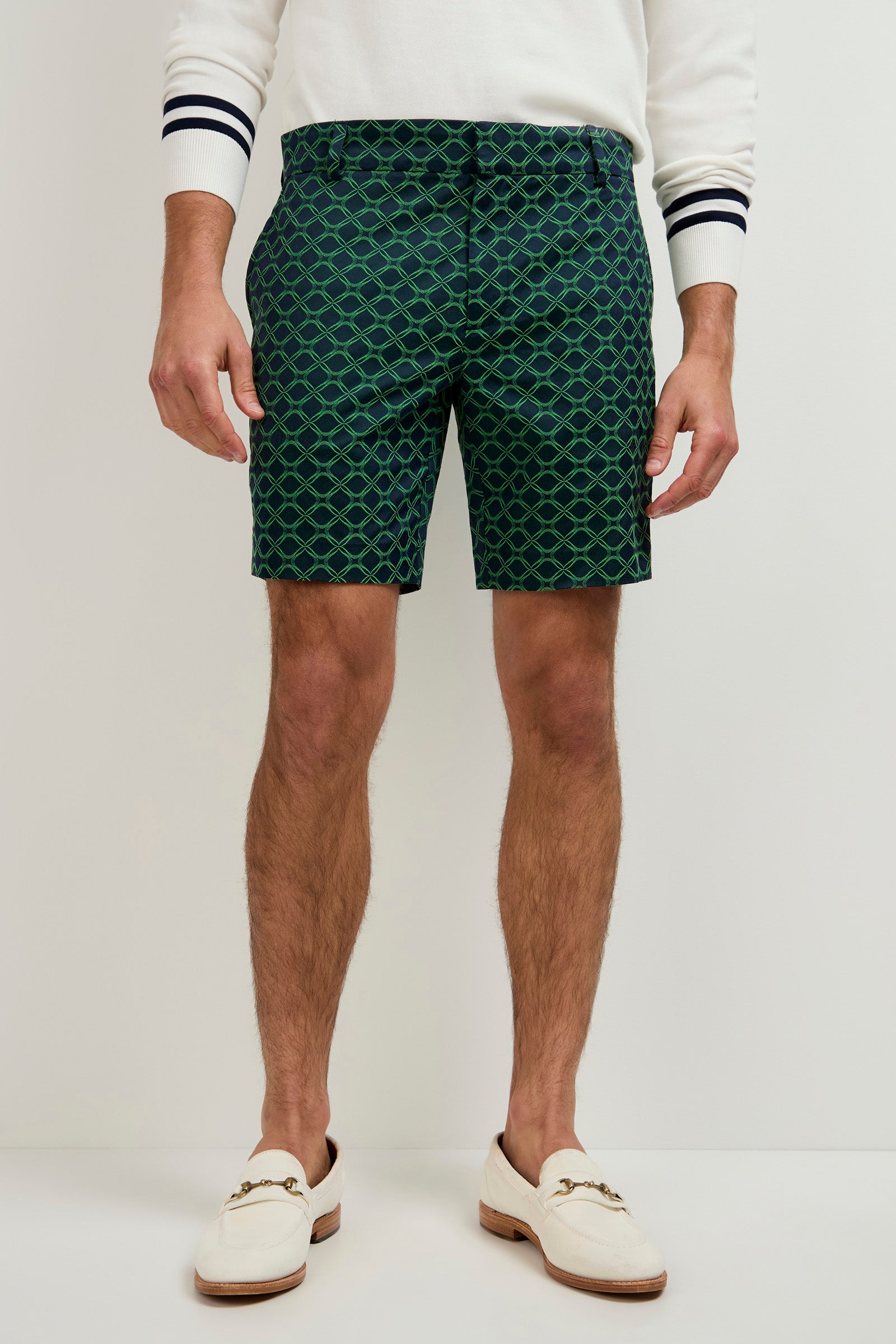 A man stands against a plain background wearing the Lawrence Short - Clover Geo Jacquard, a white long-sleeve shirt with black-striped cuffs, and white loafers with gold detailing. His hands are relaxed by his sides.