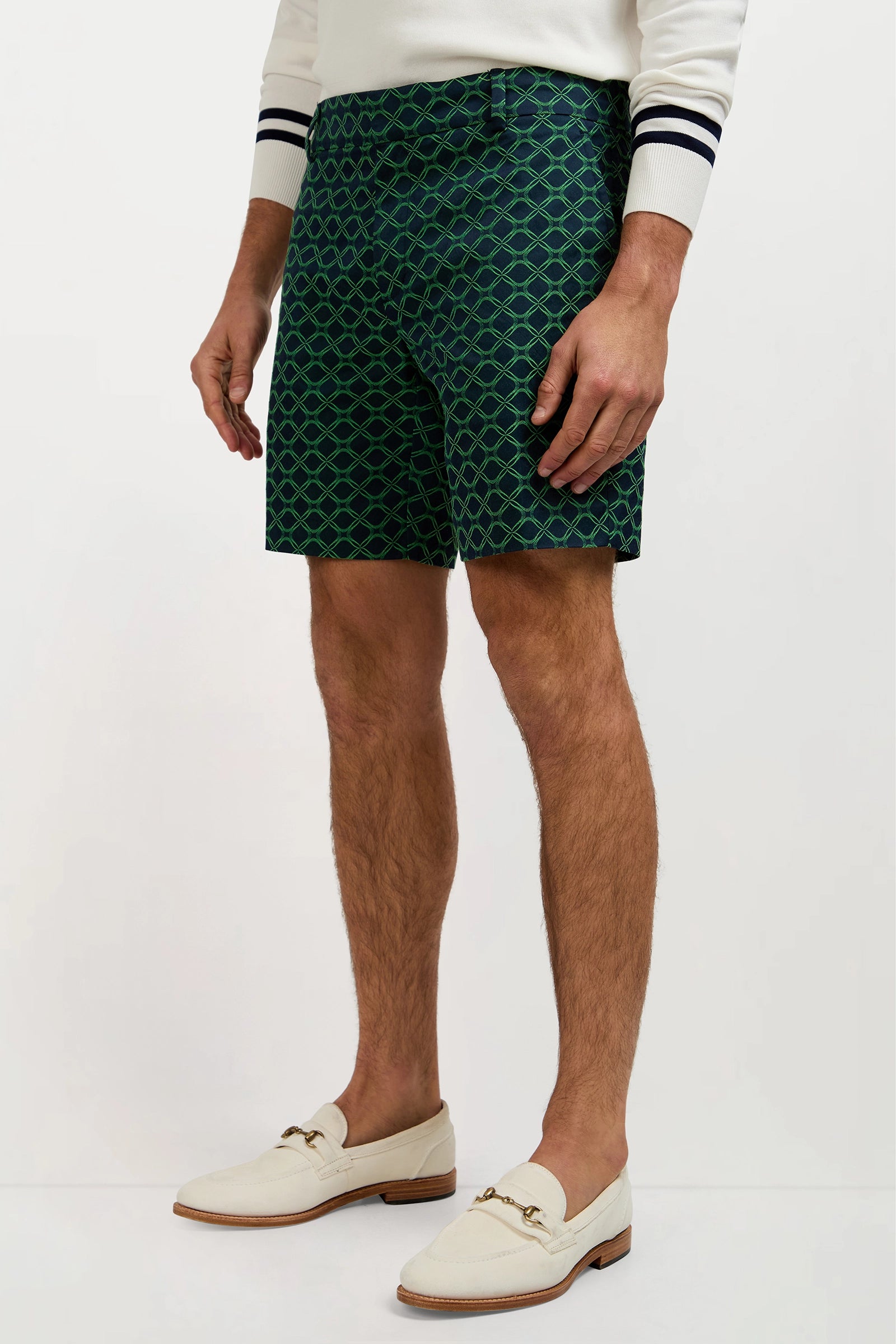 A man models the Lawrence Short - Clover Geo Jacquard, paired with a white sweater with navy sleeve stripes and cream loafers. His body is shown from shoulders down against a plain background.