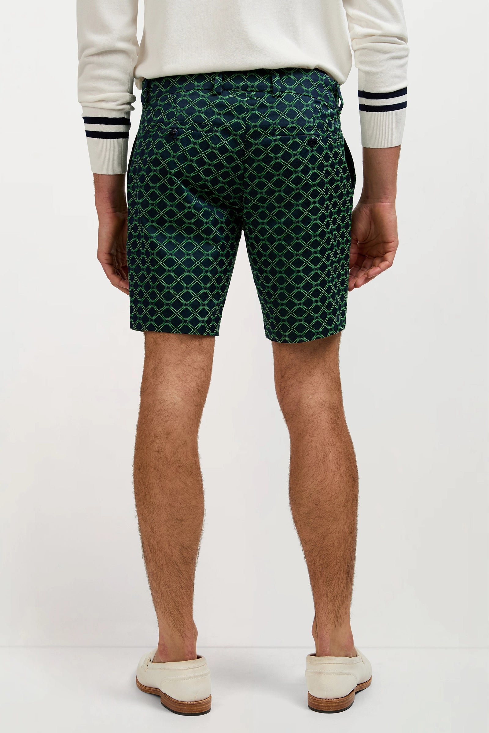 A man stands facing away, wearing the Lawrence Short – Clover Geo Jacquard, a white long-sleeve shirt with dark striped cuffs, and light-colored loafers. Only his body from shoulders to calves is visible.