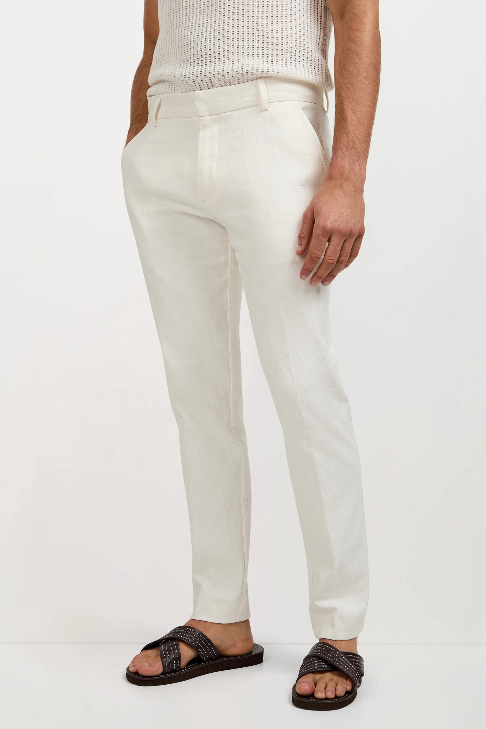 A person wearing the Clyde Slim Trouser in Tegola Textured Jacquard, a sleeveless knit top, and black sandals stands with hands in pockets against a plain background.