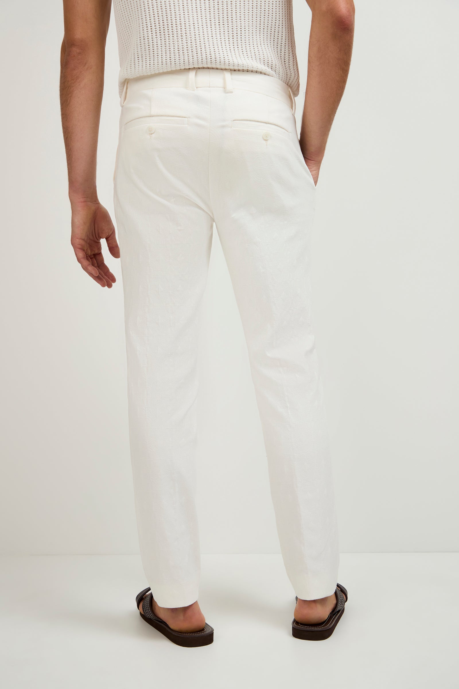 A person faces away against a plain light background, wearing the Clyde Slim Trouser in Tegola Textured Jacquard paired with a white sleeveless knit top and dark sandals.