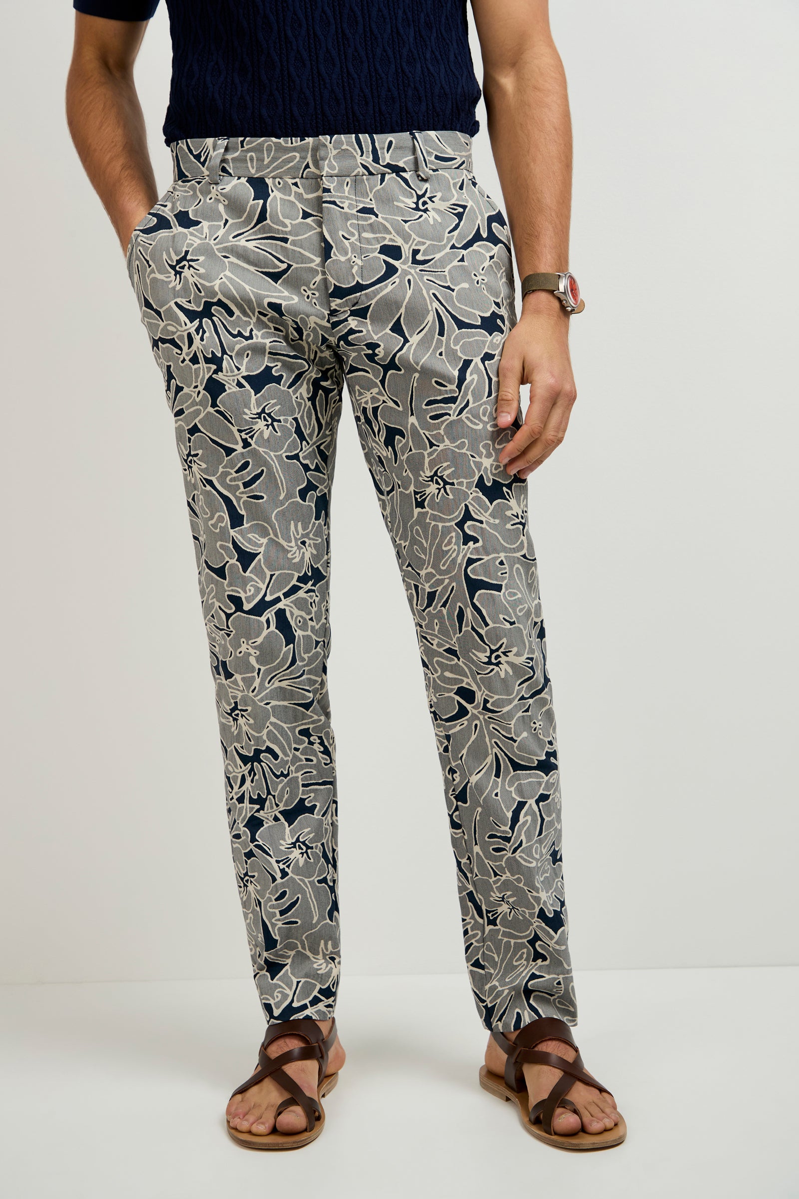 A person stands against a plain background, wearing the Clyde Slim Trouser in Artsy Floral Jacquard with a dark short-sleeve shirt, brown sandals, and a watch. Their hands are in their pockets.