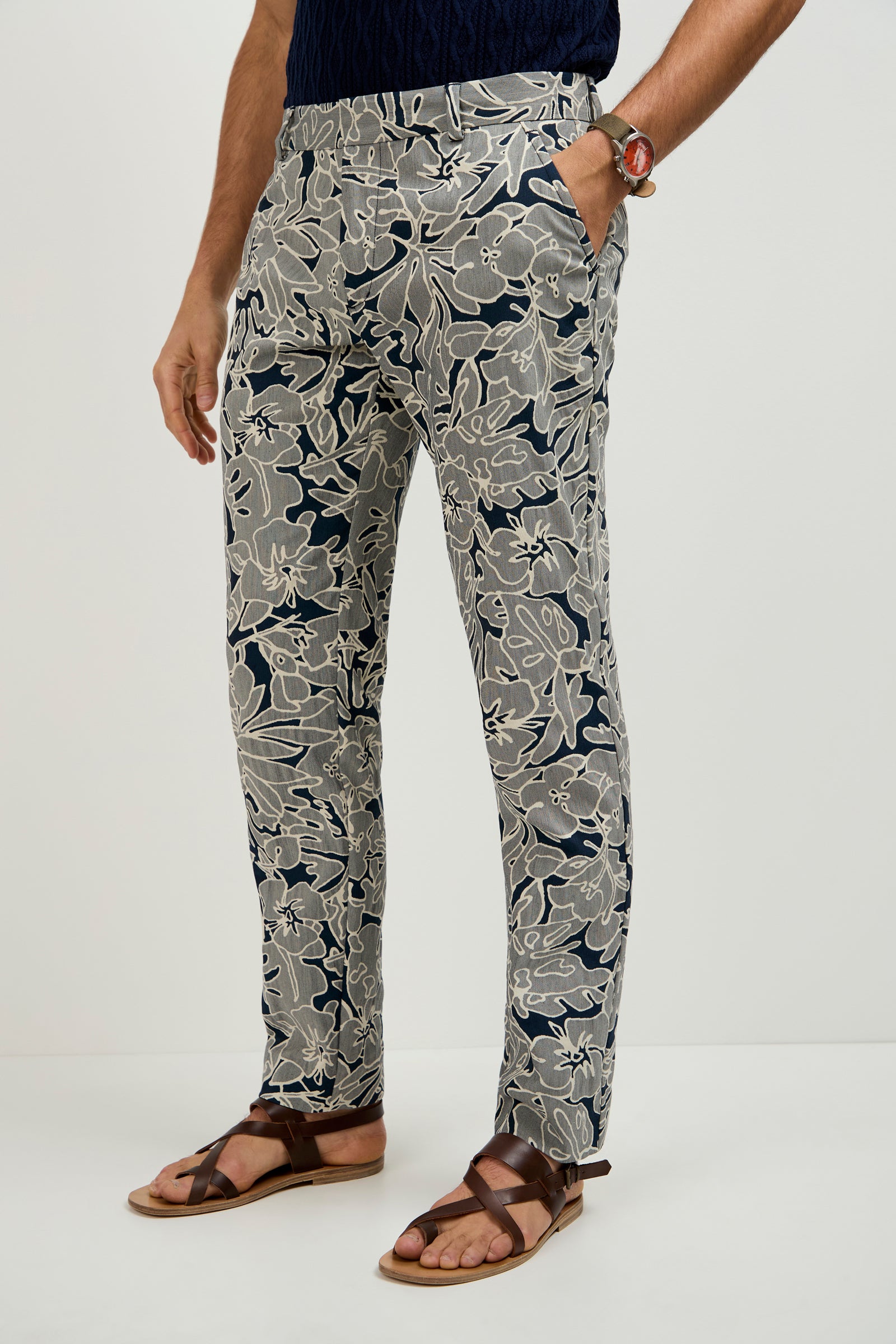 A person wearing the Clyde Slim Trouser - Artsy Floral Jacquard, paired with a dark blue shirt, brown leather sandals, and a wristwatch, stands against a plain light background.