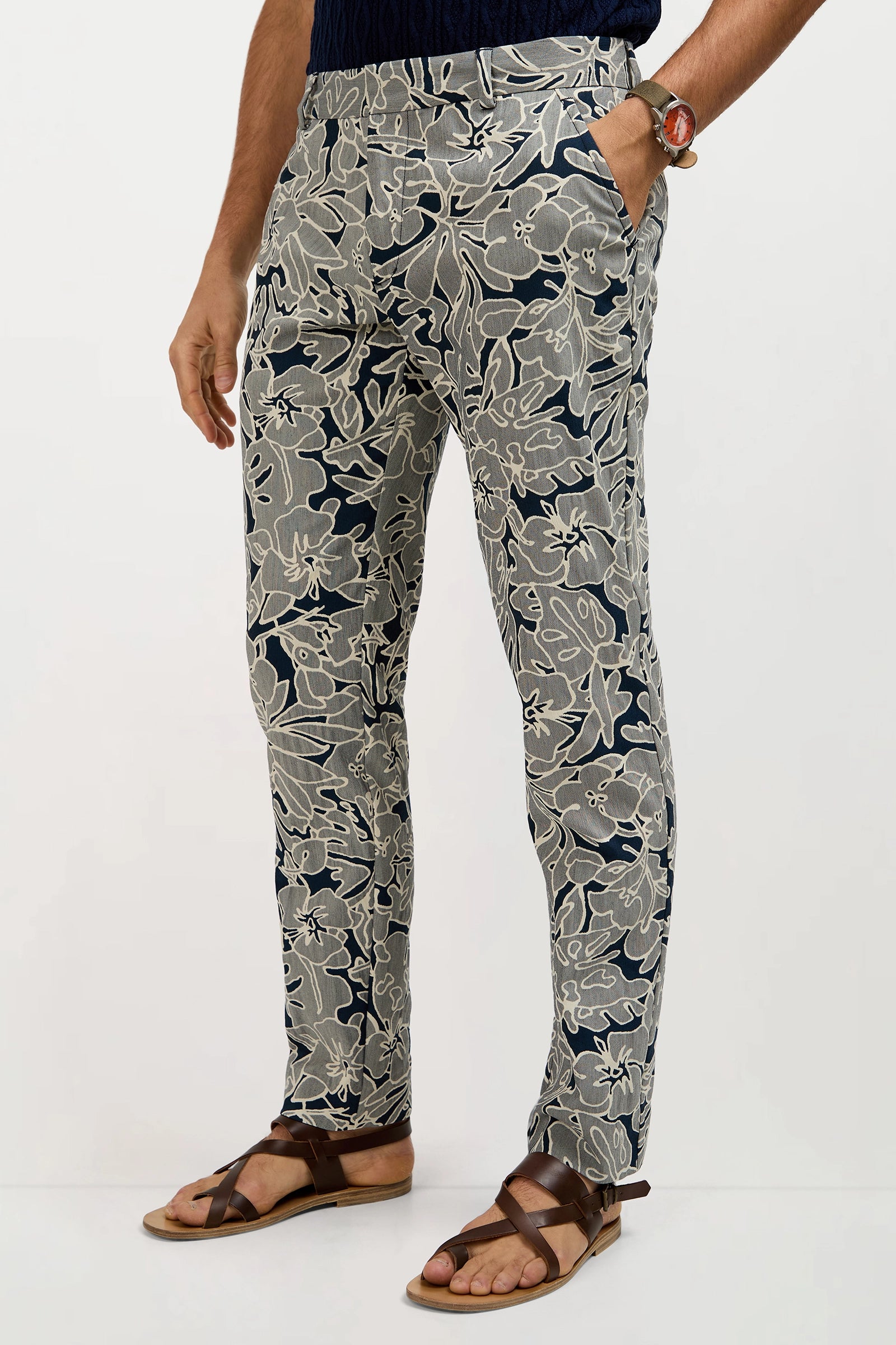 A man wears the Clyde Slim Trouser in Artsy Floral Jacquard, featuring a bold navy and beige floral pattern. He pairs them with brown sandals, a dark shirt, and a beaded bracelet on his left wrist, hands in pockets.