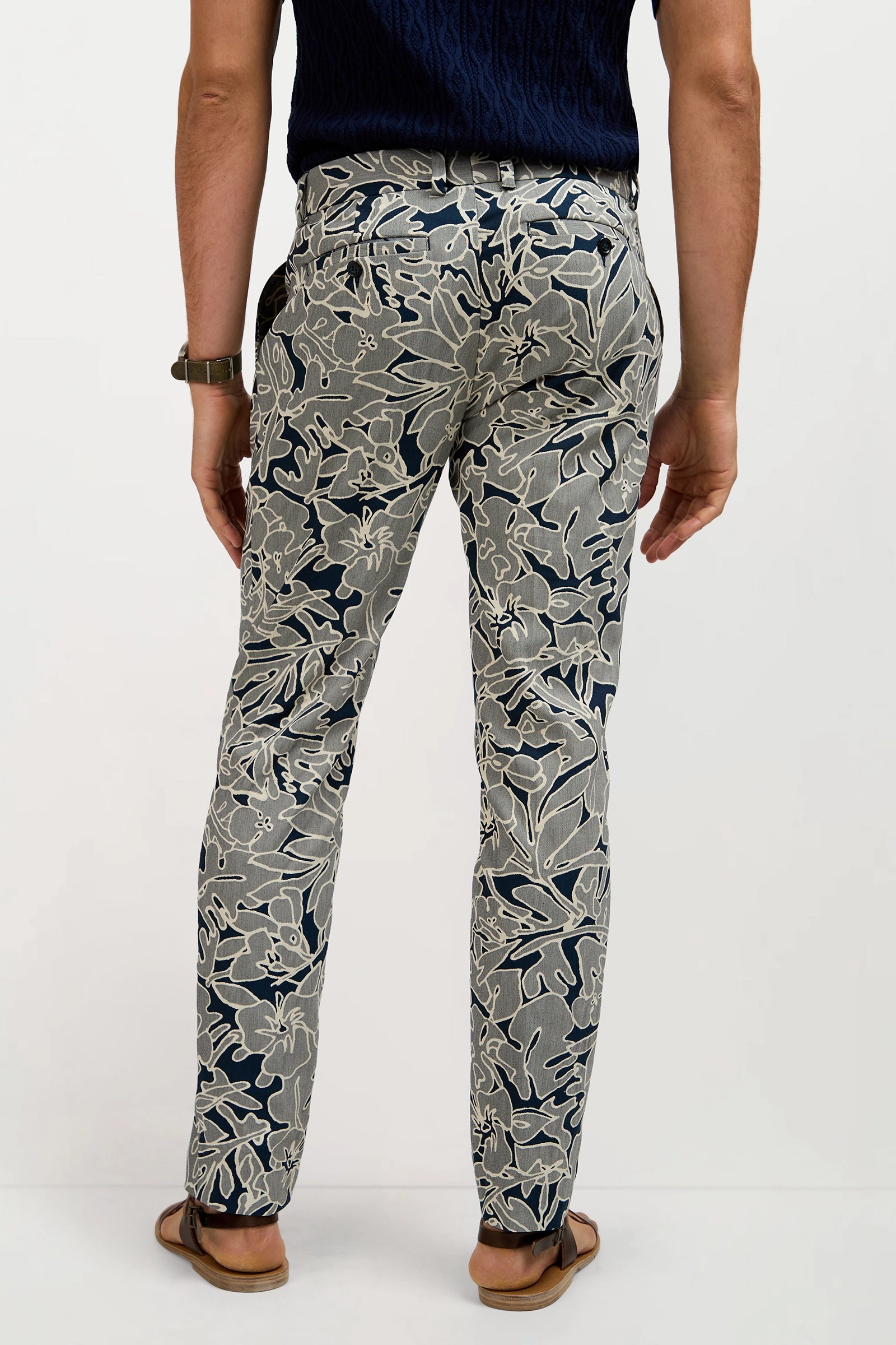 A man stands facing away, wearing Clyde Slim Trouser - Artsy Floral Jacquard with a navy knit shirt, brown watch, and brown sandals against a plain background.