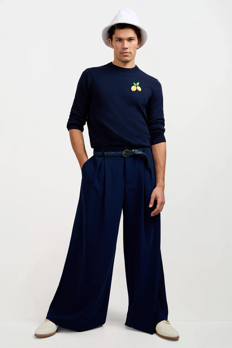 A man stands with hands in pockets, wearing a white bucket hat, a navy blue sweatshirt with a lemon patch, Clyde Wide-Leg Trouser in Ink, and white shoes against a plain background.