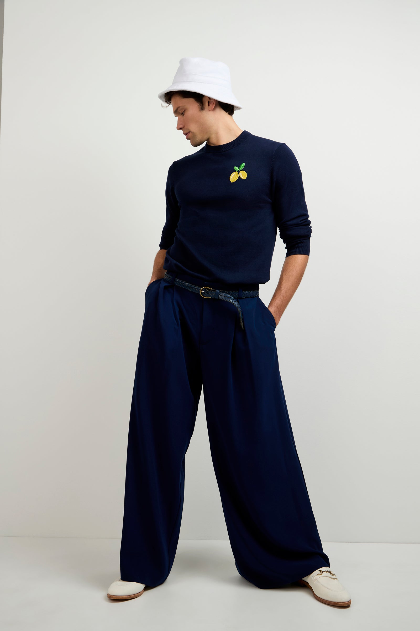 A man stands against a plain white background wearing ink-colored Clyde Wide-Leg Trousers, a navy sweater with a small fruit graphic, a white bucket hat, black belt, and white shoes.