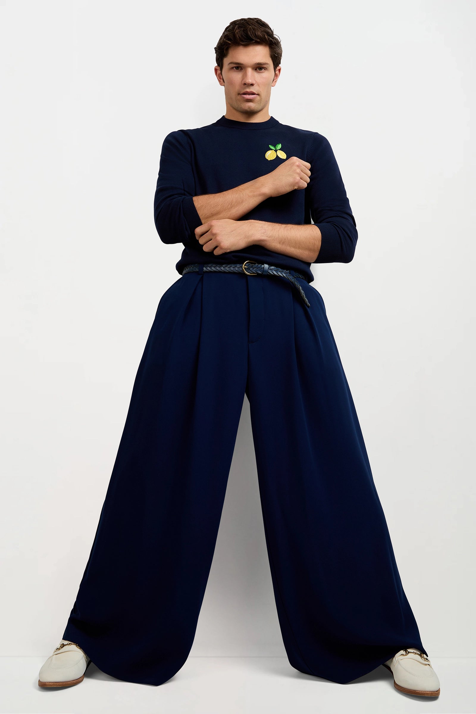 A man stands against a plain white background with arms crossed, wearing navy wide-legged Clyde Wide-Leg Trousers, a navy sweater featuring a lemon graphic, a braided belt, and white shoes in an assertive pose.