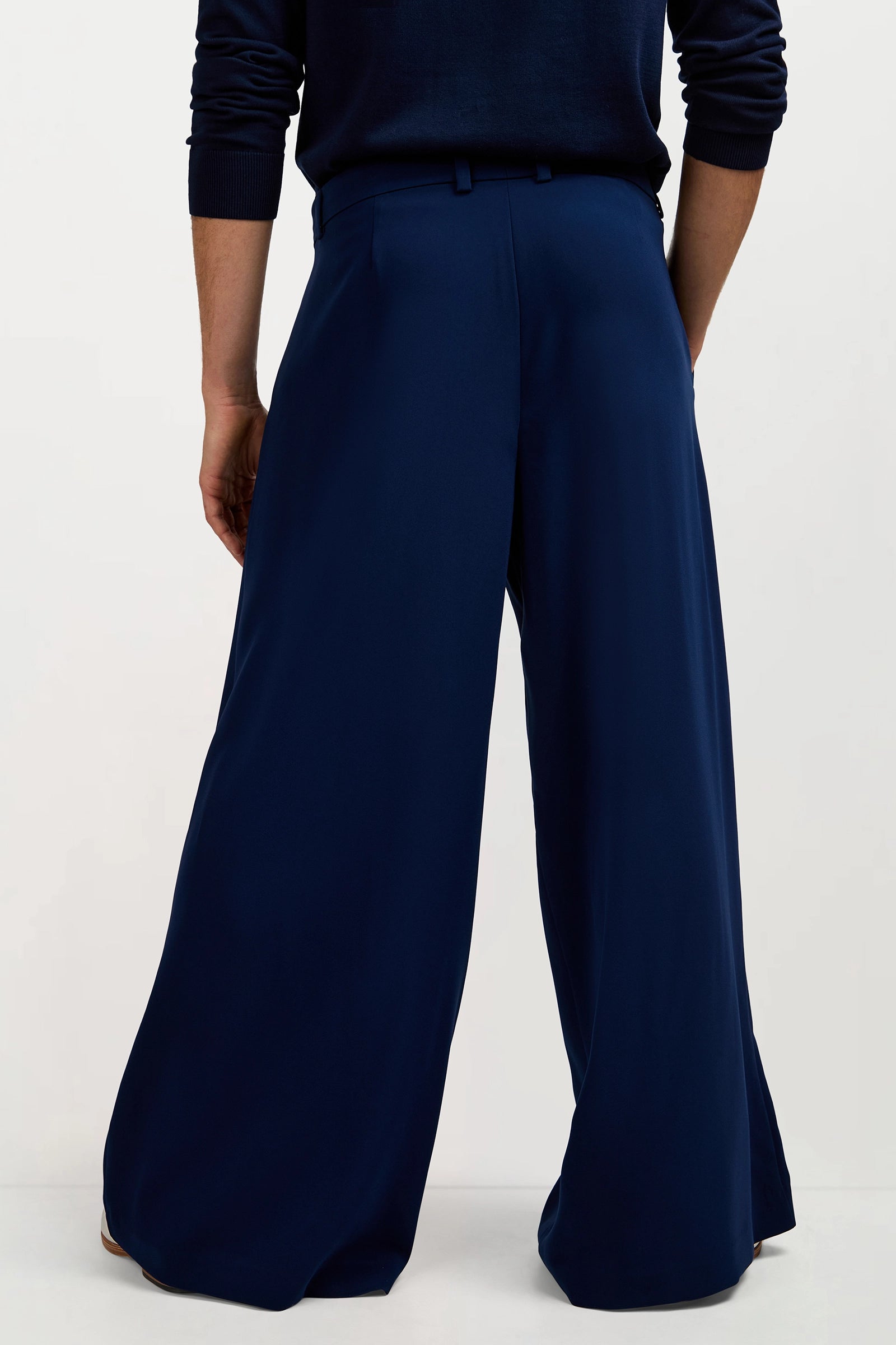 Someone stands with their back to the camera in a plain light setting, wearing the Clyde Wide-Leg Trouser in navy blue paired with a matching dark long-sleeve top.