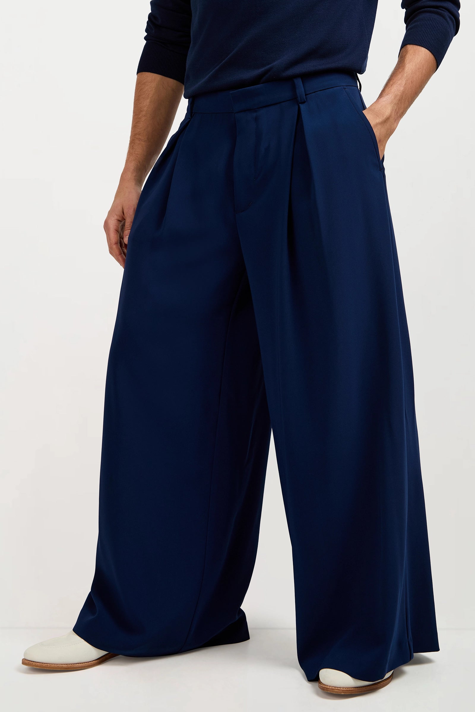 Someone stands against a white background with hands in pockets, wearing the Clyde Wide-Leg Trouser in navy blue, paired with a matching dark long-sleeve top and white shoes.
