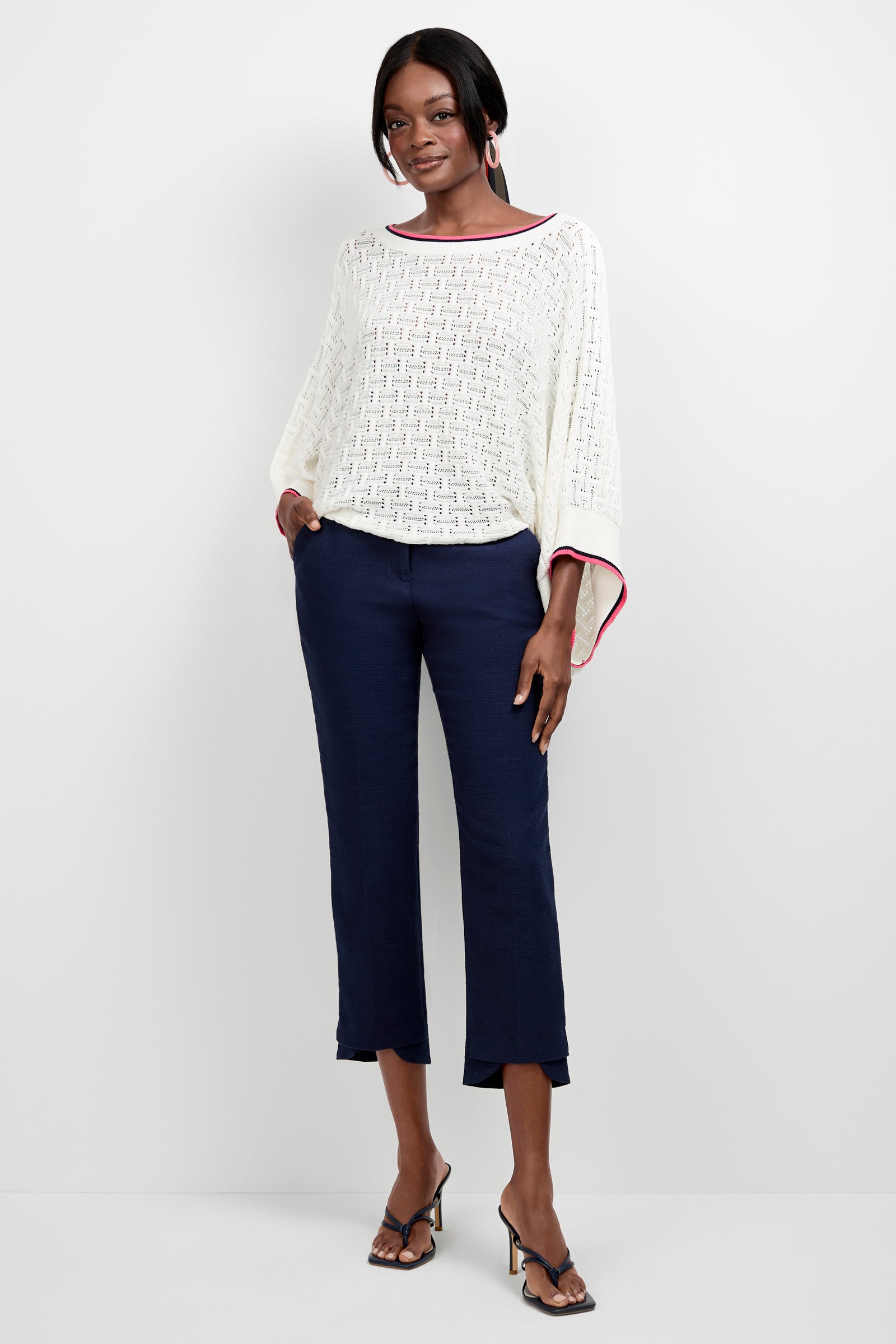 A woman with short hair smiles with her hands in her pockets, wearing a white textured blouse, black heeled sandals, and navy Ore Button-Hem Cropped Pants against a white background.