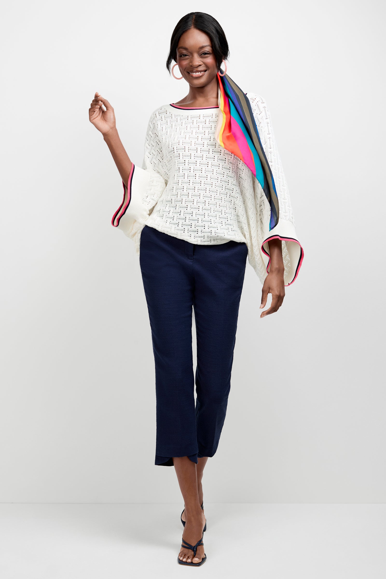 A woman smiles against a plain white background, wearing the Horizon Boatneck Pullover—a white textured top with wide sleeves—paired with navy cropped pants, black sandals, and a colorful scarf tied in her hair.