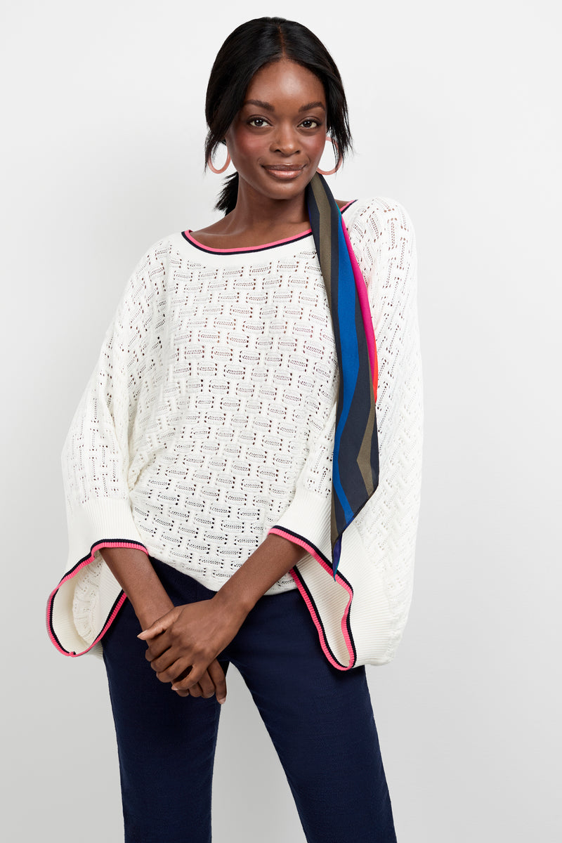 A woman stands against a white background, smiling softly with her hands clasped, wearing the Horizon Boatneck Pullover—a white knit sweater with red trim—paired with navy pants and a colorful scarf in her hair.