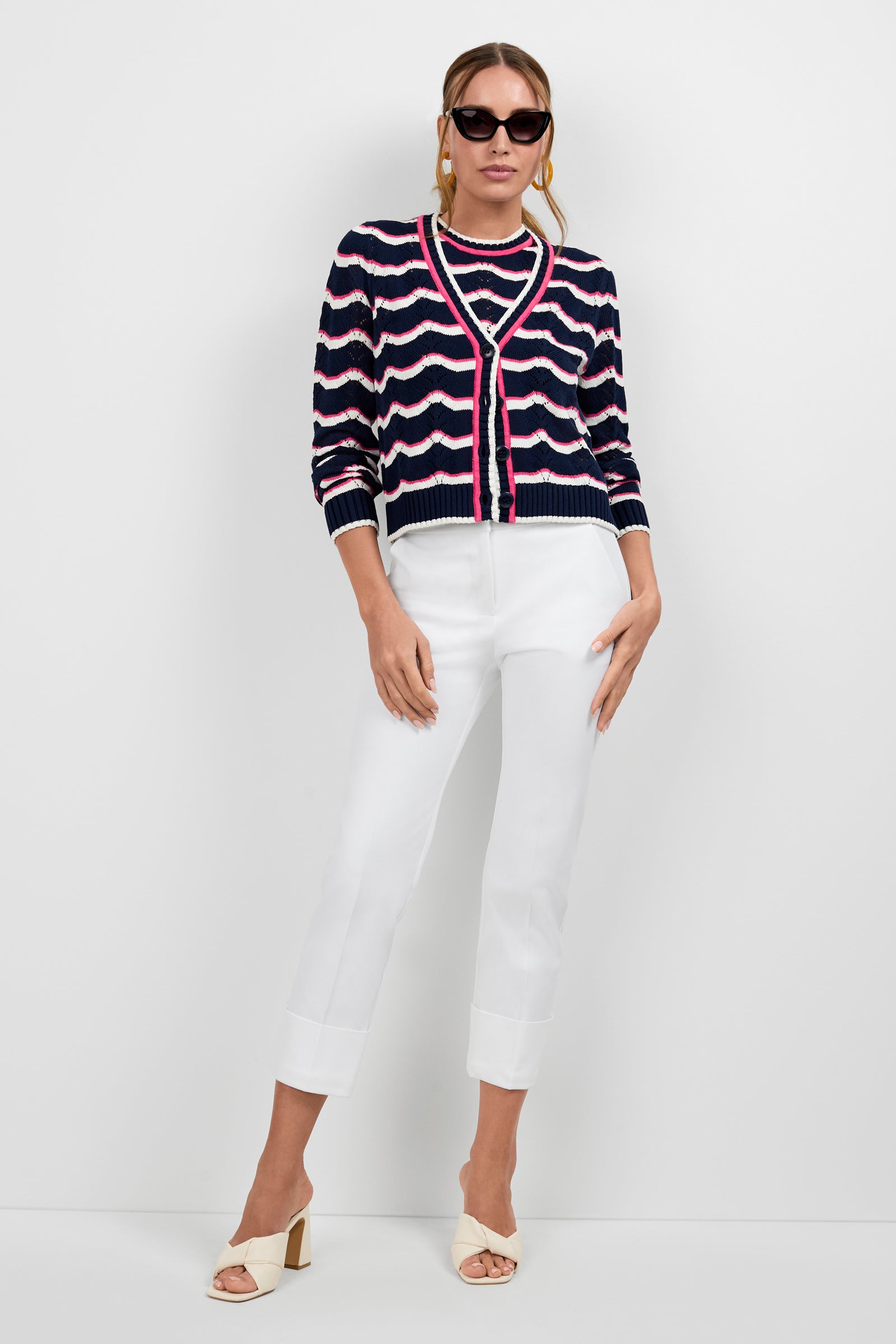 A woman poses against a white wall wearing sunglasses, the navy blue, pink, and white wave-patterned Alessia Knit Cardigan over a matching top, white cuffed pants, and beige open-toe block-heeled sandals.
