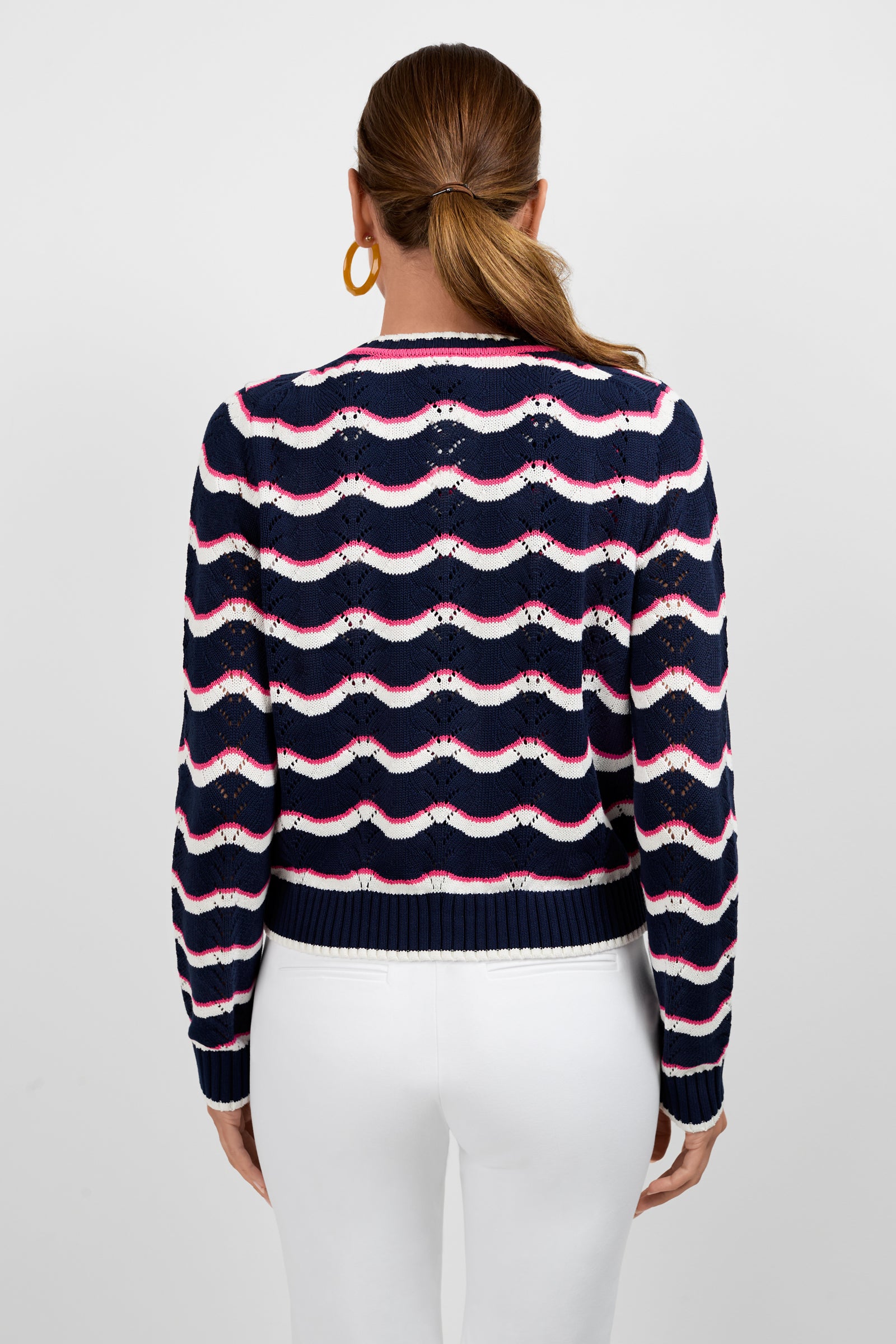 A woman with brown hair in a low ponytail is seen from the back, wearing the Alessia Knit Cardigan in navy blue with white and pink wavy stripes, paired with white pants and large gold hoop earrings against a plain light background.
